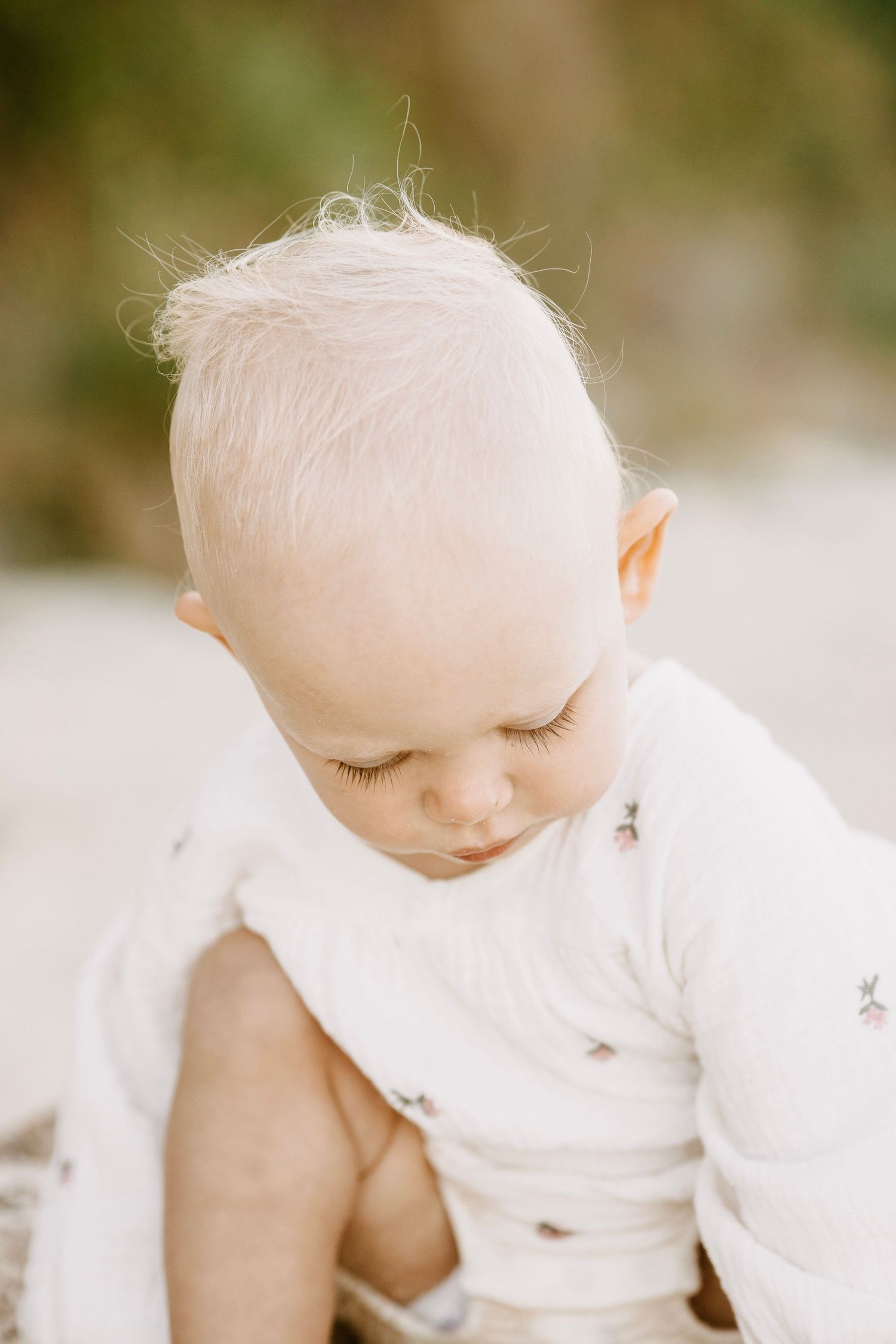 Close-up portrait of a peaceful baby with fine blonde hair wearing a white floral romper outdoors.