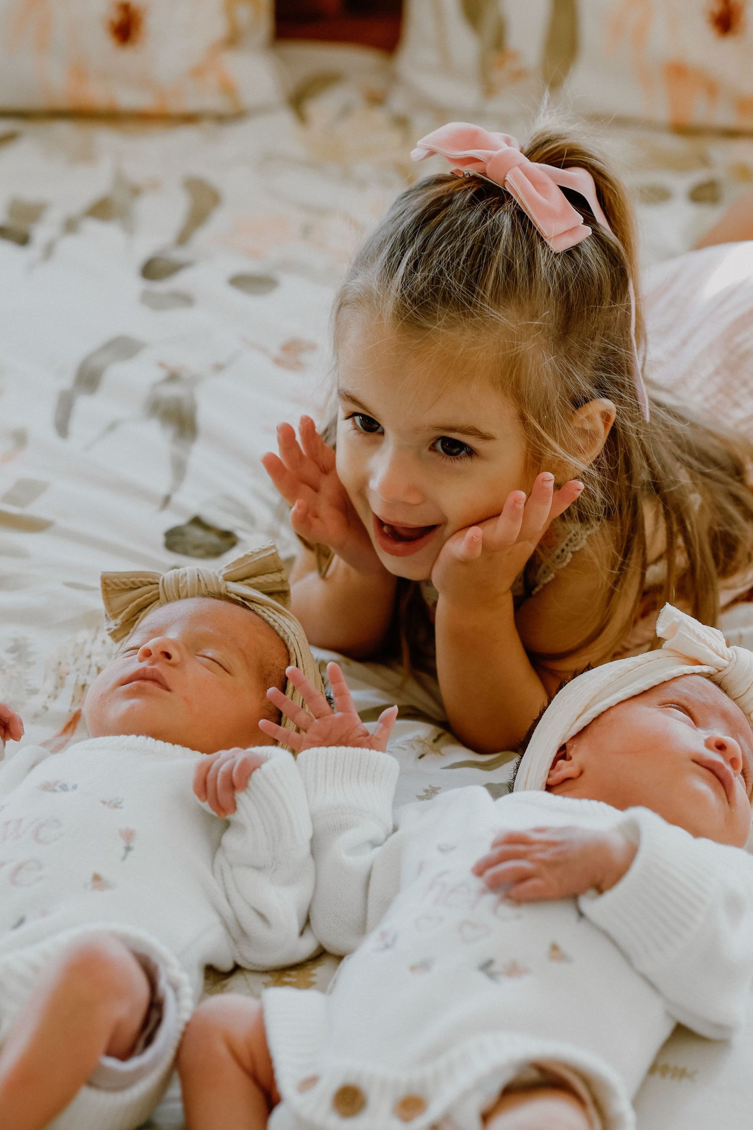 Excited toddler girl with pink bow admiring two sleeping newborn twins on a floral bedspread.