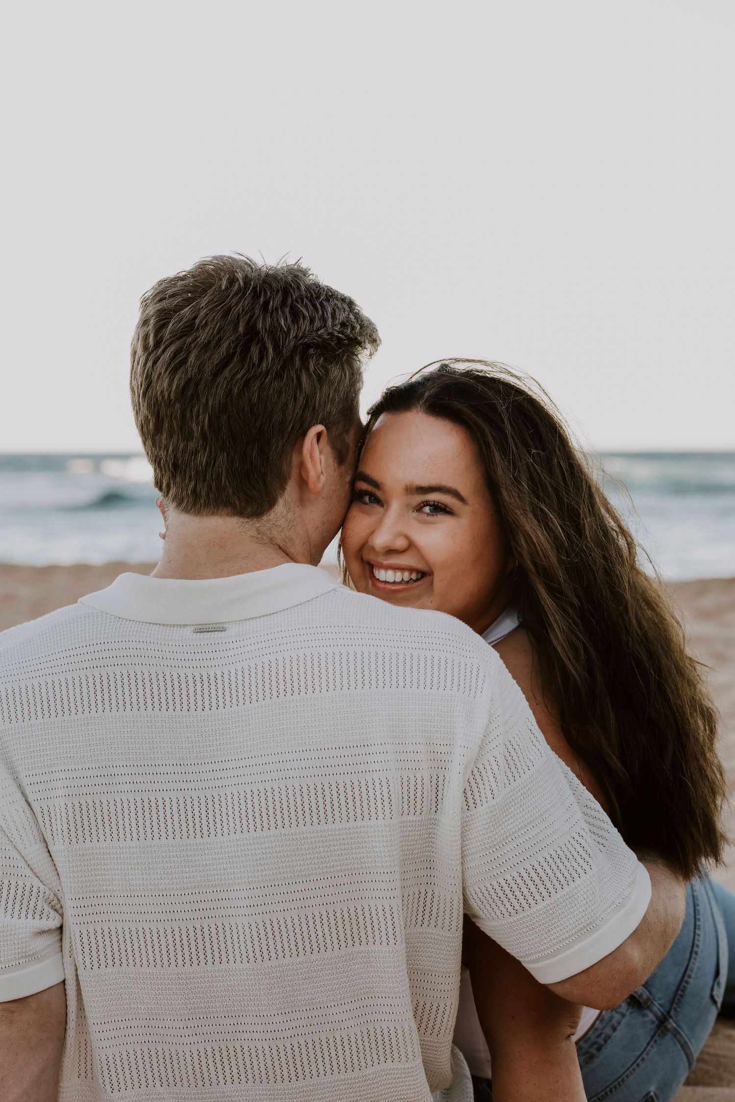 Happy woman smiling over mans shoulder during romantic beach photo session at sunset.