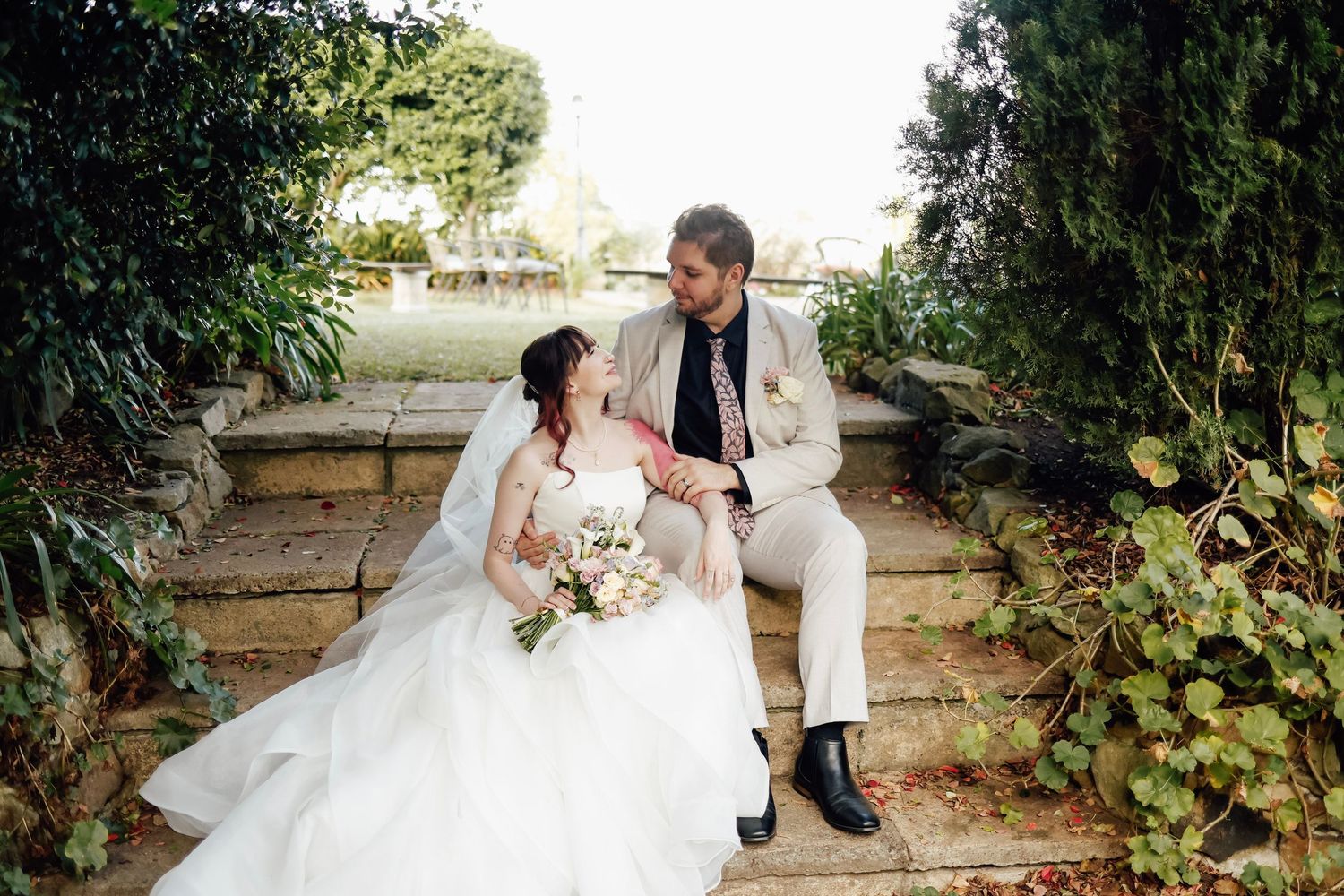 Bride and groom sitting on stone steps in a lush garden, sharing a tender moment on their wedding day.