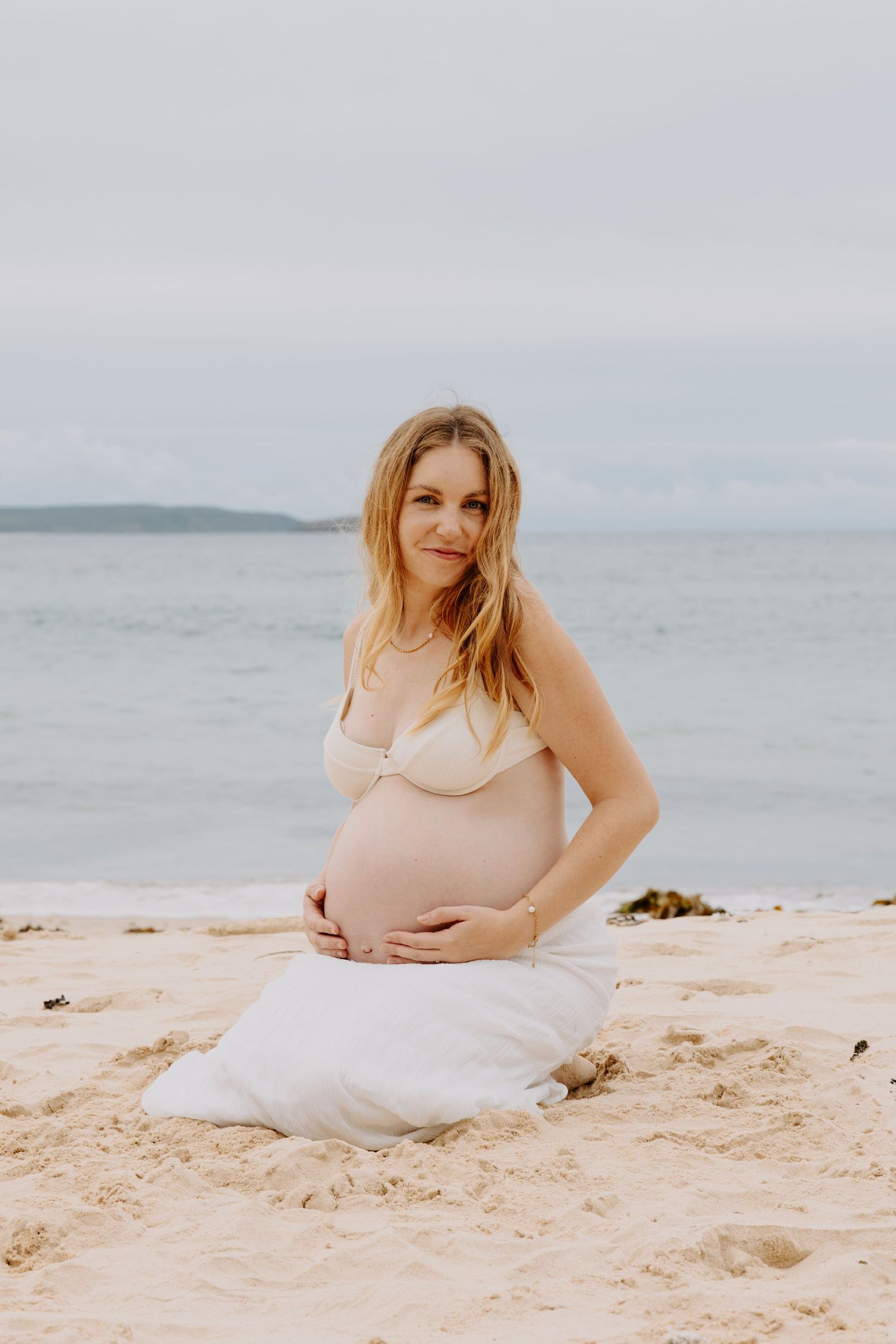 Pregnant woman in beige bikini top kneeling on beach sand cradling baby bump by ocean.
