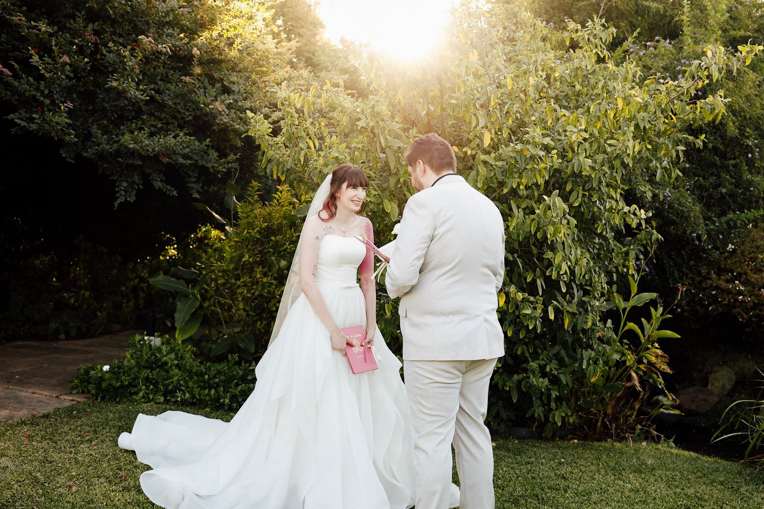 Bride in white gown and groom in tan suit share a tender moment in a sunlit garden.