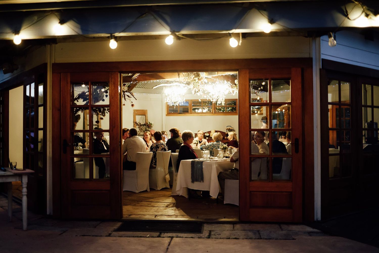 Elegant restaurant dining room viewed through open French doors at night, with guests seated at white-clothed tables under a chandelier.