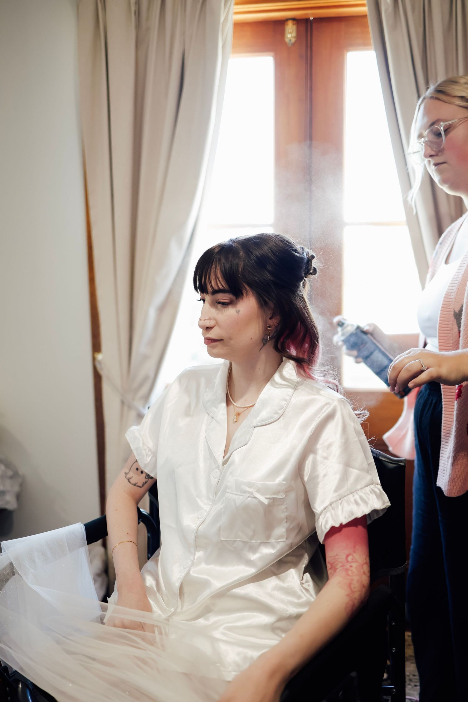 Bride getting hair styled by hairdresser while sitting in white robe before wedding ceremony.