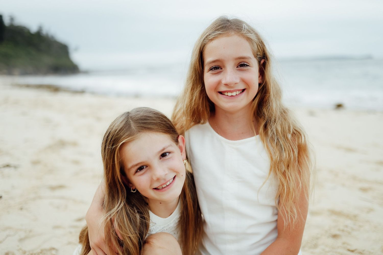 Two smiling young girls with long blonde hair hugging on a sandy beach.