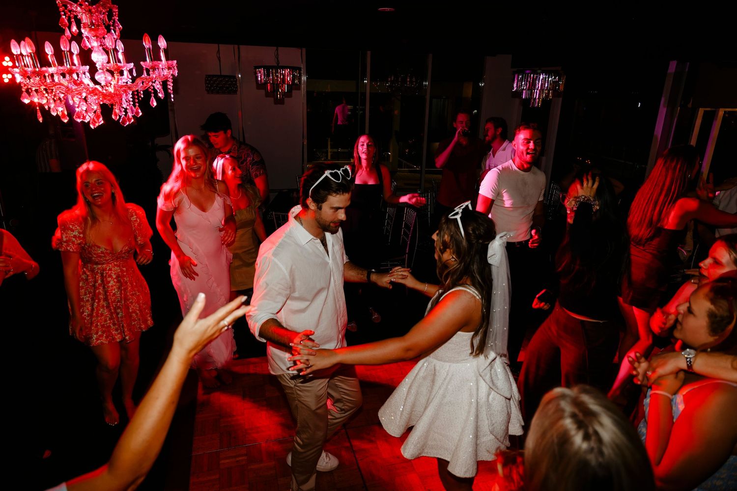 Bride and groom dancing surrounded by celebrating guests under red lights at wedding reception.