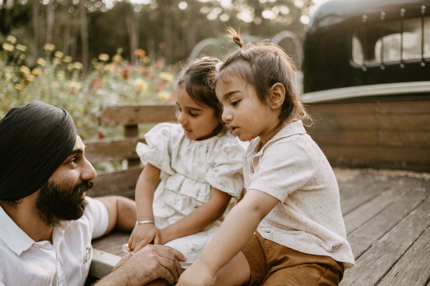 A Sikh father sits on a wooden deck with two young daughters in a sunlit garden setting.