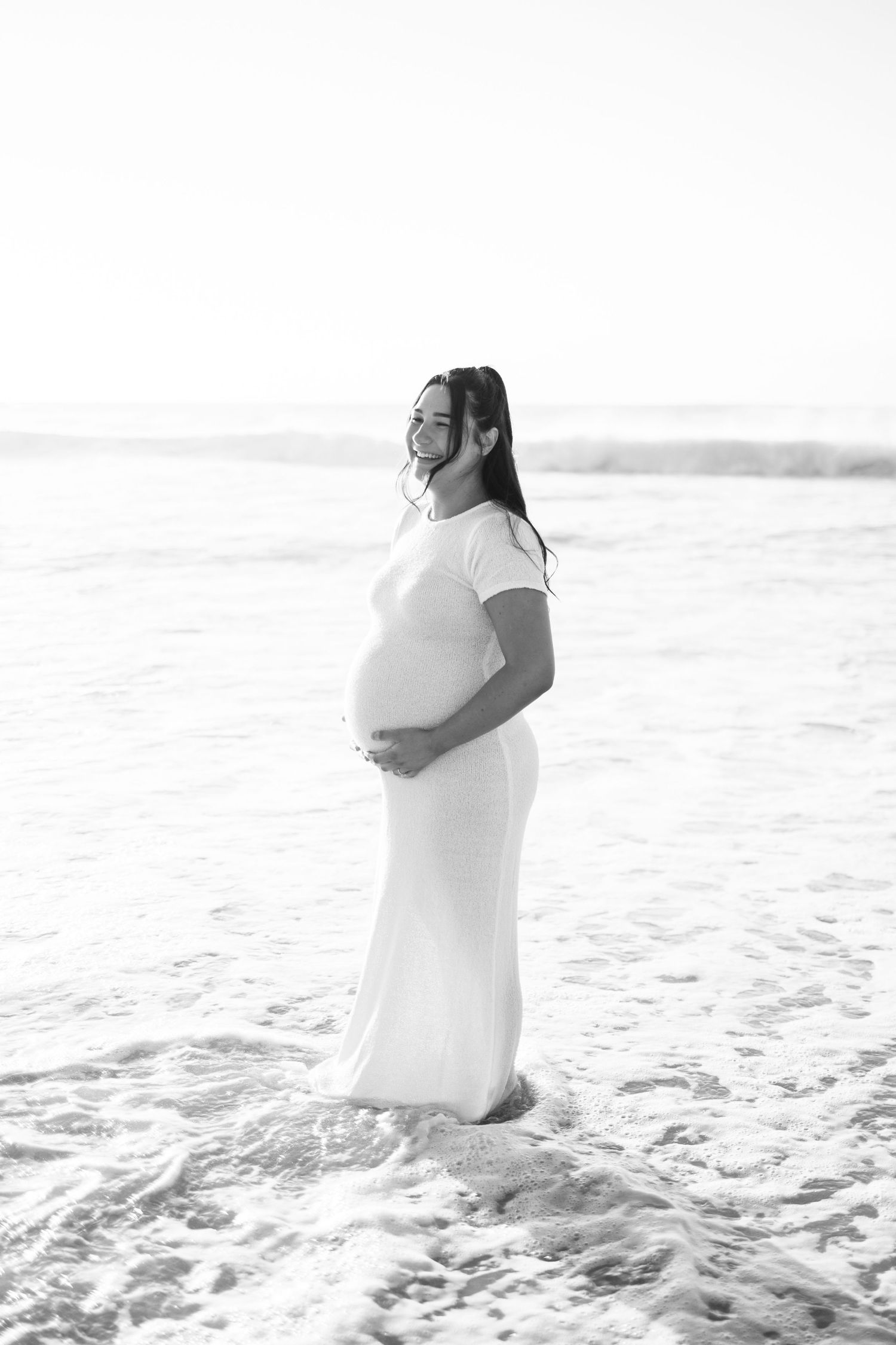 Black and white photo of pregnant woman in white dress standing in ocean waves.