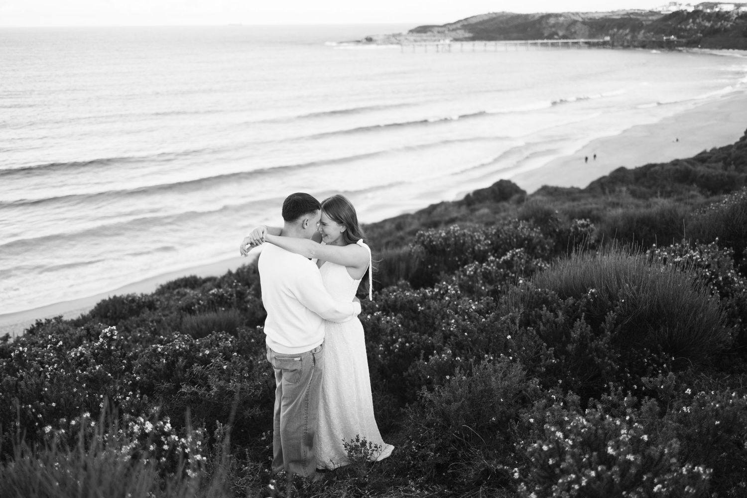 Black and white photo of a couple embracing on a coastal hillside with ocean and beach in the background.