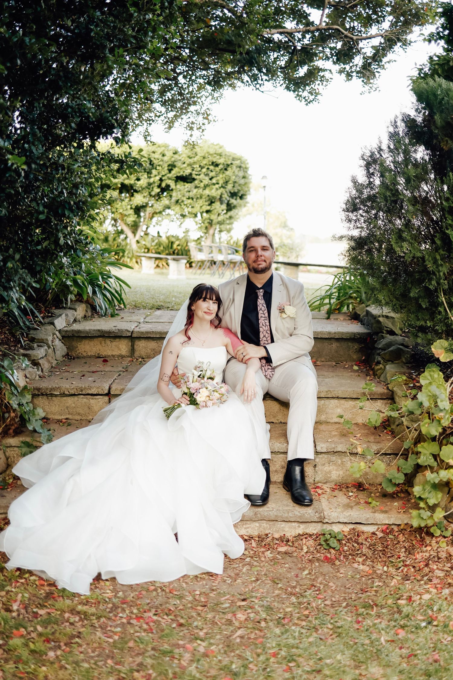 Bride and groom seated on stone steps in lush garden, bride holding soft floral bouquet.