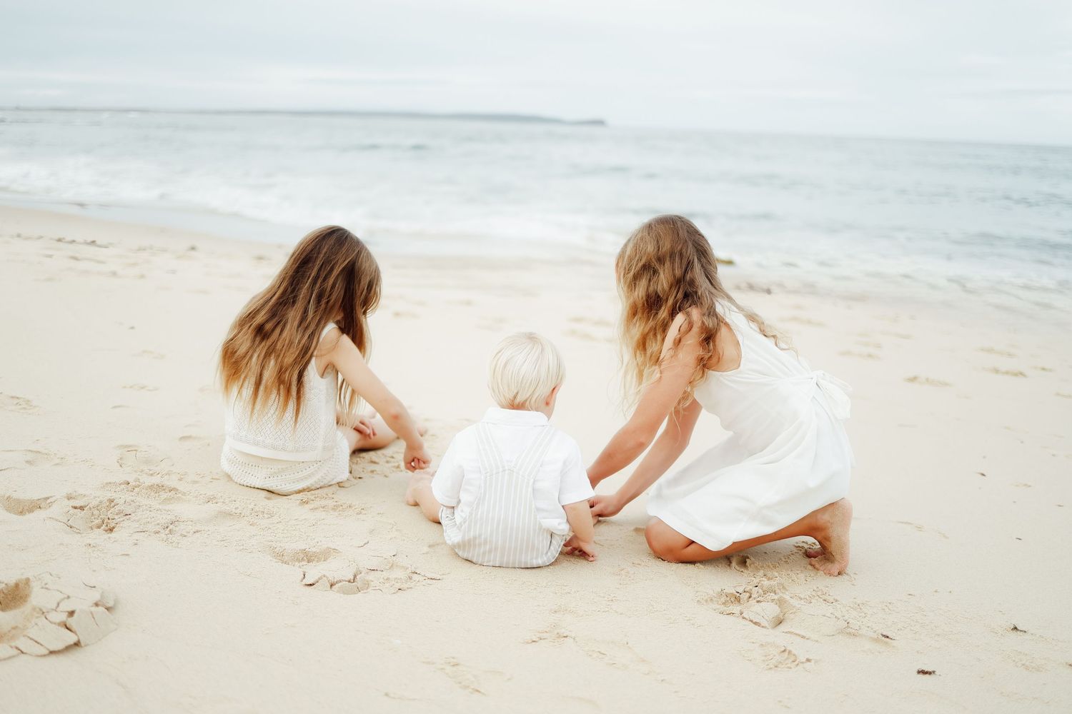 Three young children in white outfits playing together in the sand at the beach.