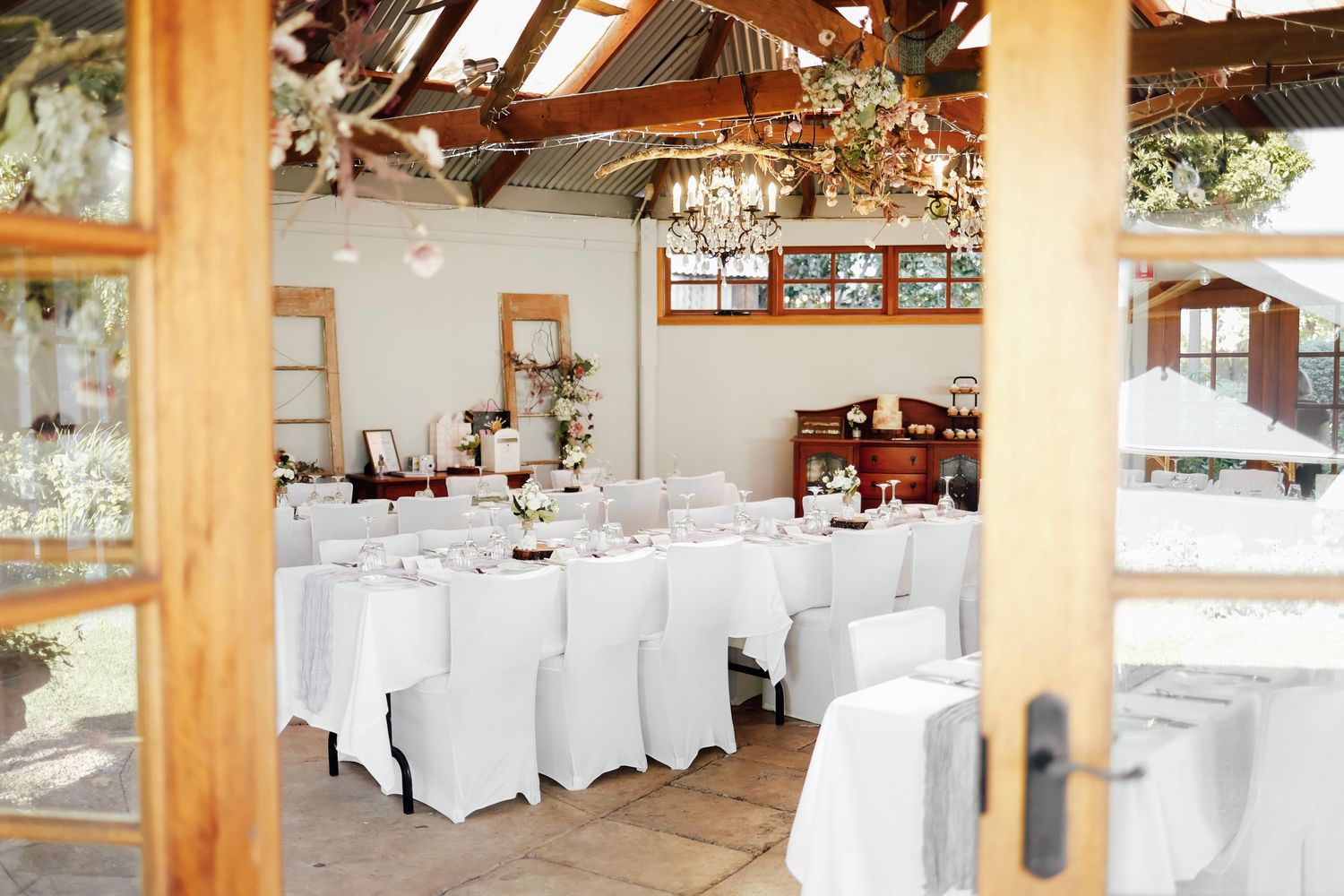 Elegant wedding reception venue with white-draped tables, wooden beams, and floral chandeliers in a bright rustic barn setting.