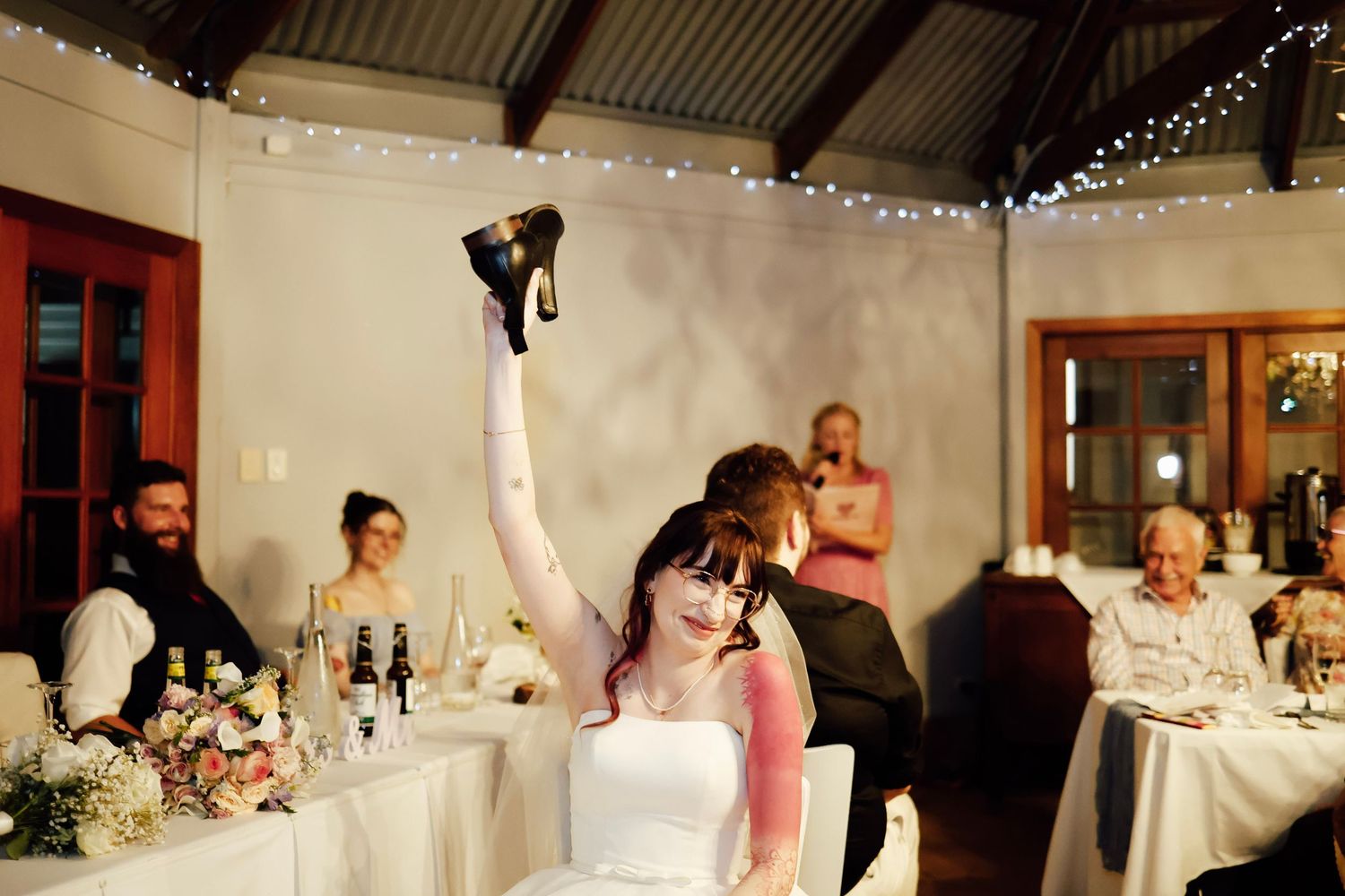 Joyful bride raises shoes in celebration during indoor wedding reception with fairy lights.