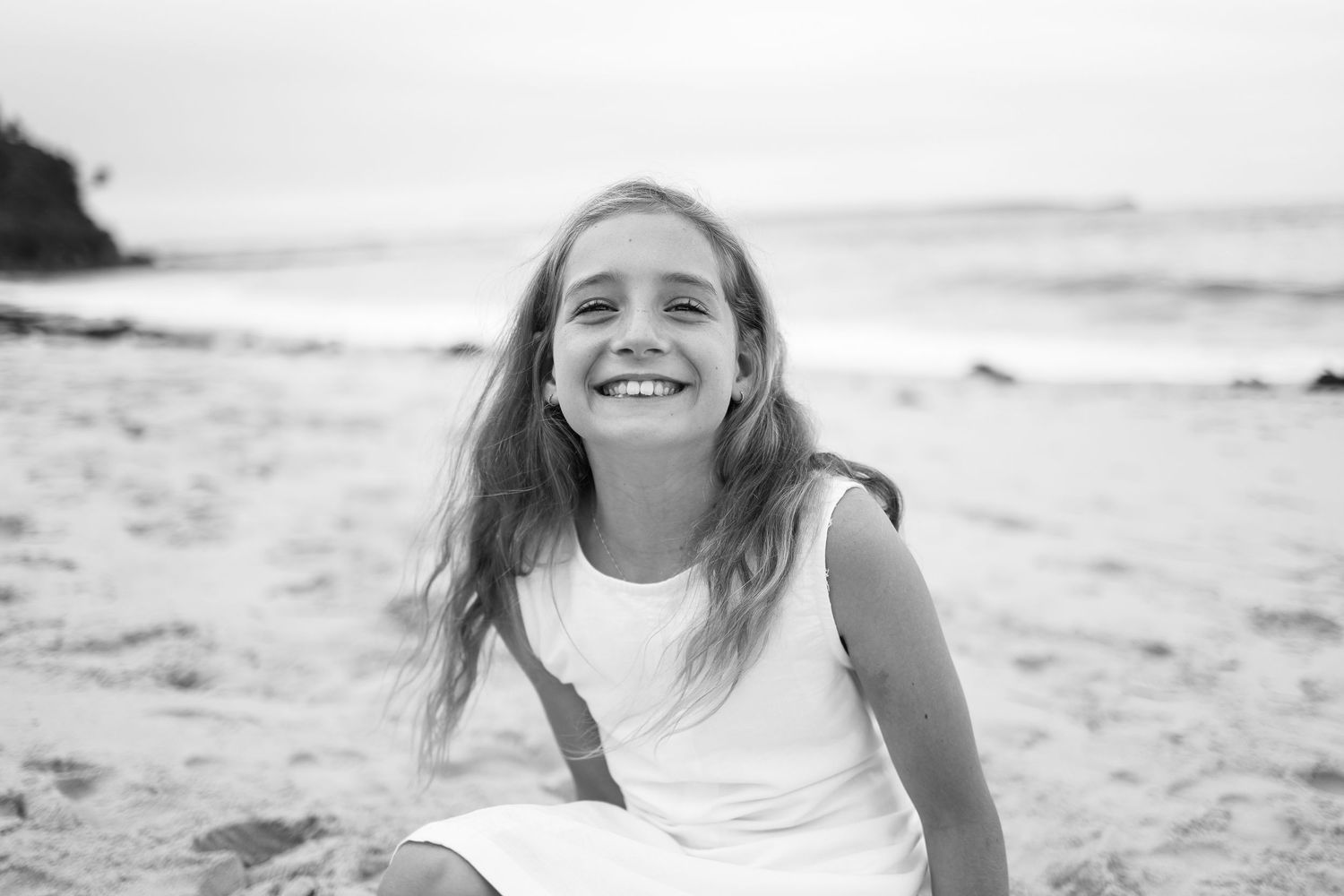 Black and white portrait of laughing girl with long hair sitting on beach in white dress.