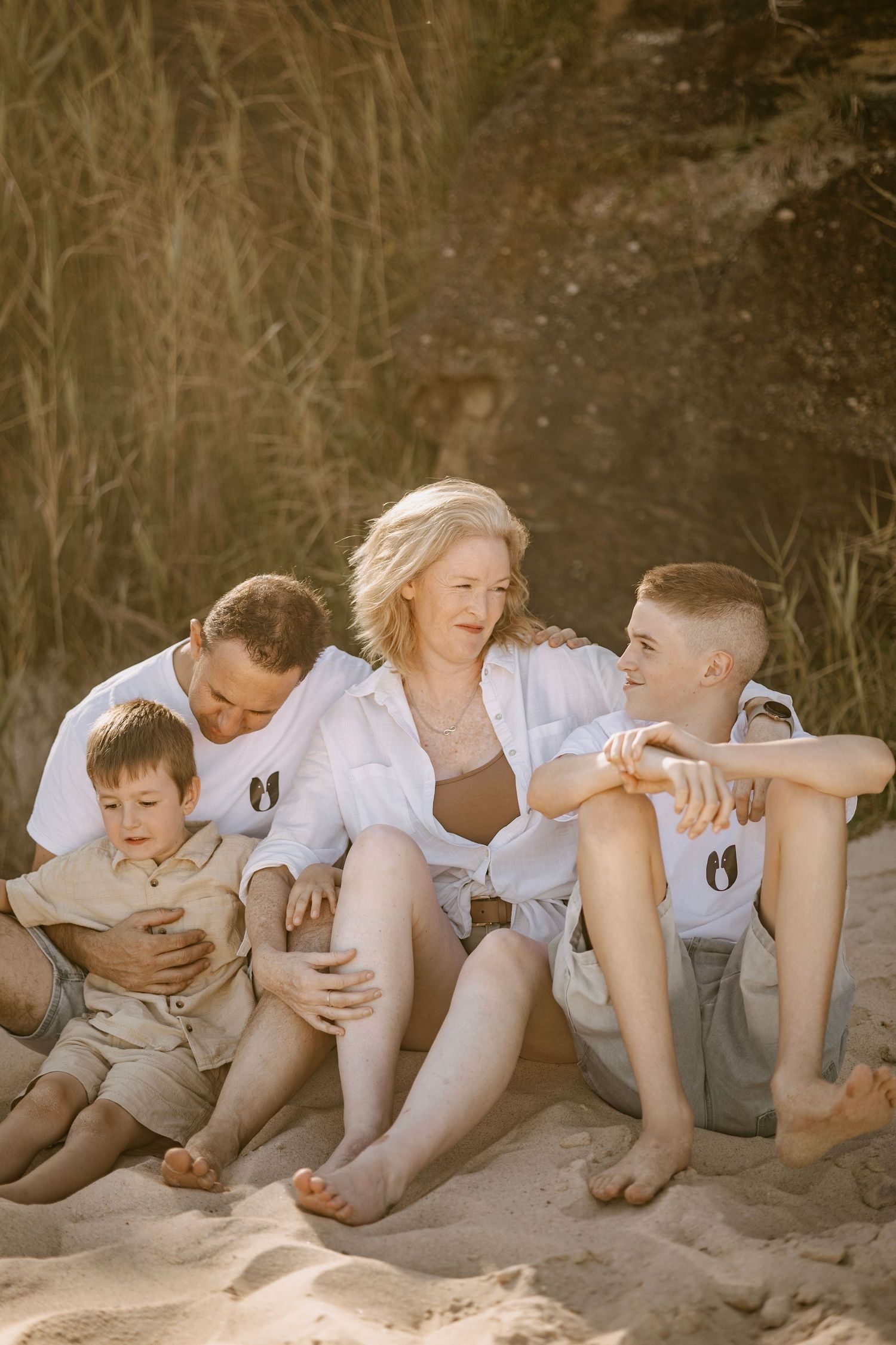 Family of four sitting together on sandy beach, parents embracing two boys in warm golden sunset light.