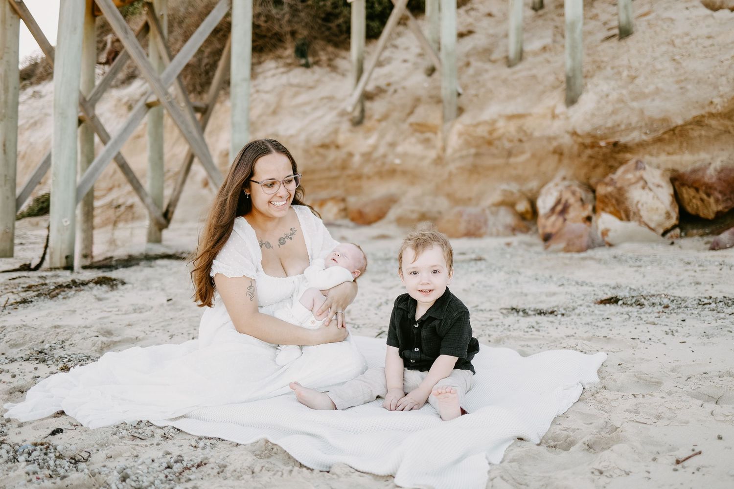 A mother sits on a white blanket on the beach holding a newborn while a toddler sits beside her.