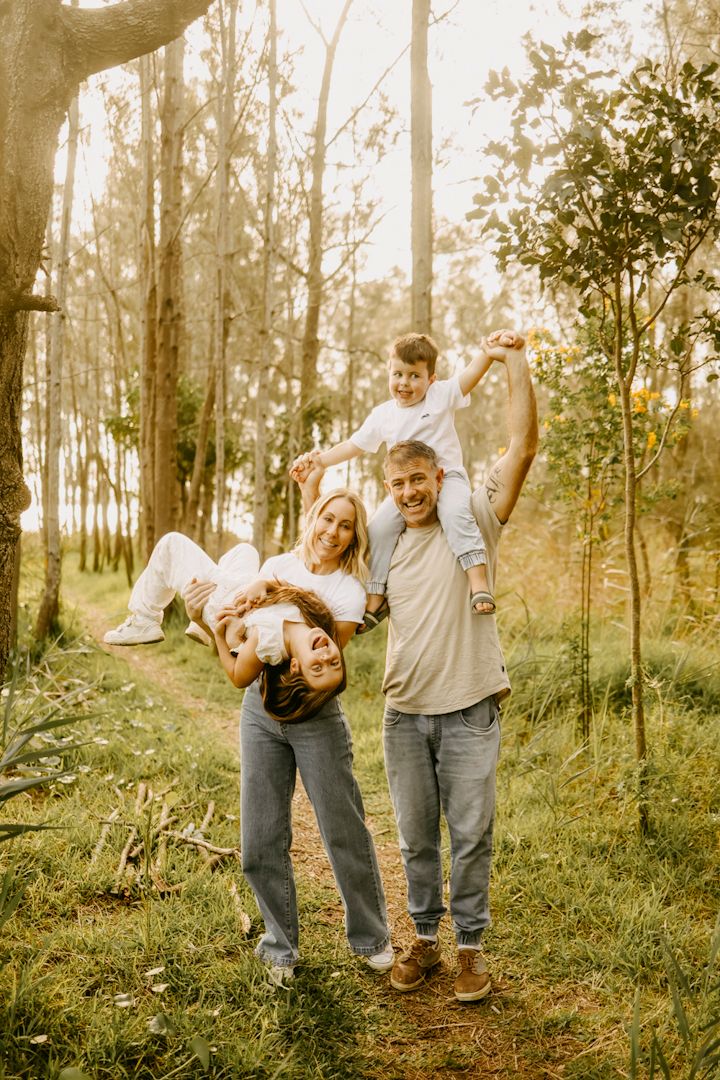 A joyful family plays in a forest, parents carrying their two laughing children on their backs.