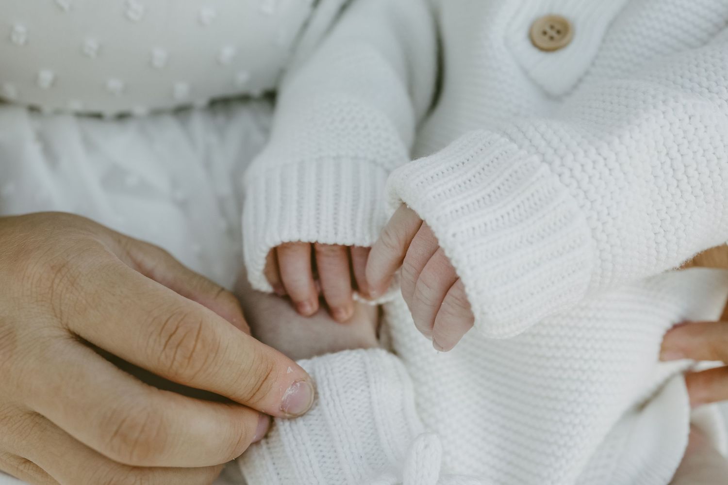 Close-up of newborn baby's tiny hand grasping parent's finger, wearing white knit sweater.