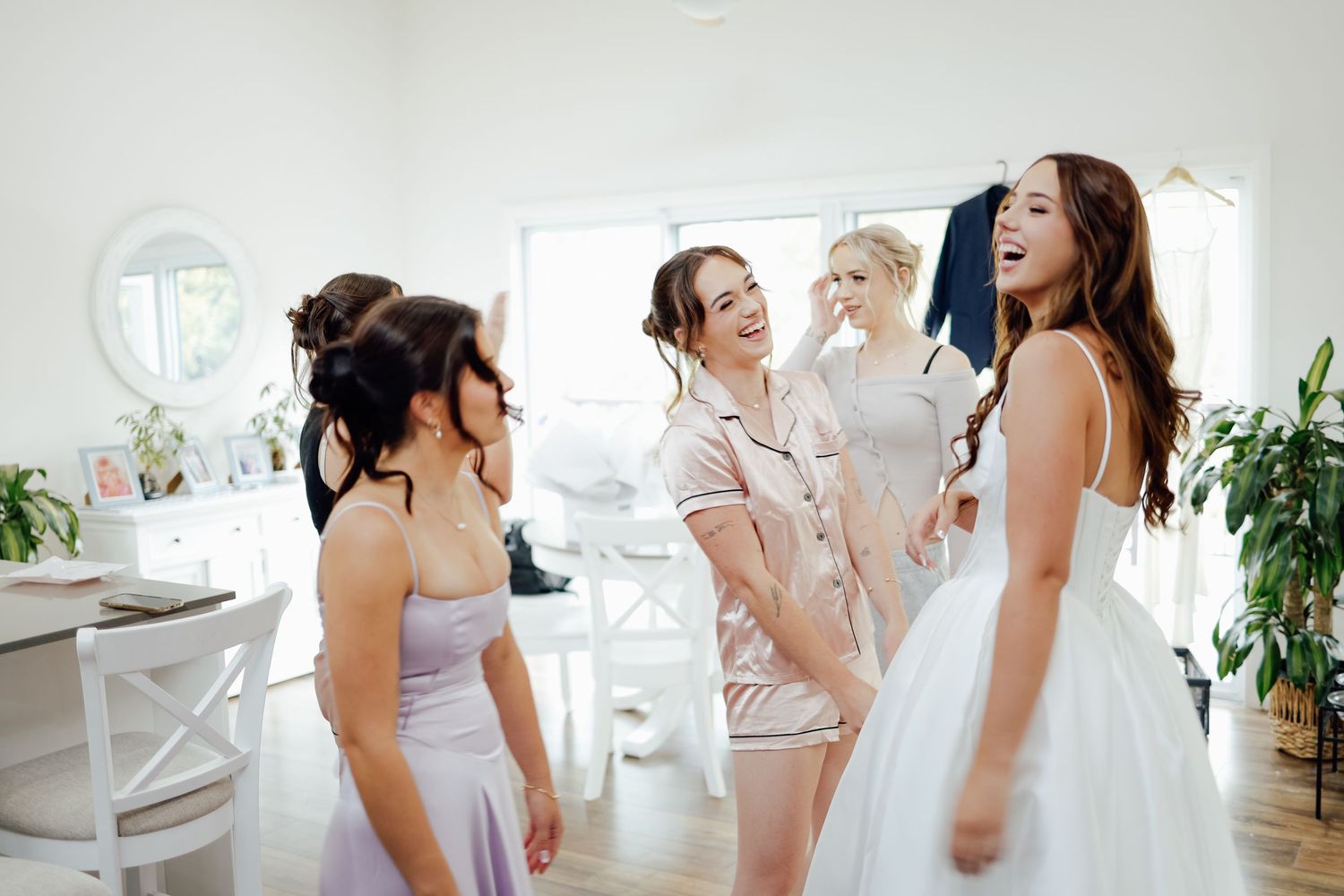 Bride in white gown laughing with bridesmaids in lavender dresses during wedding preparations indoors.