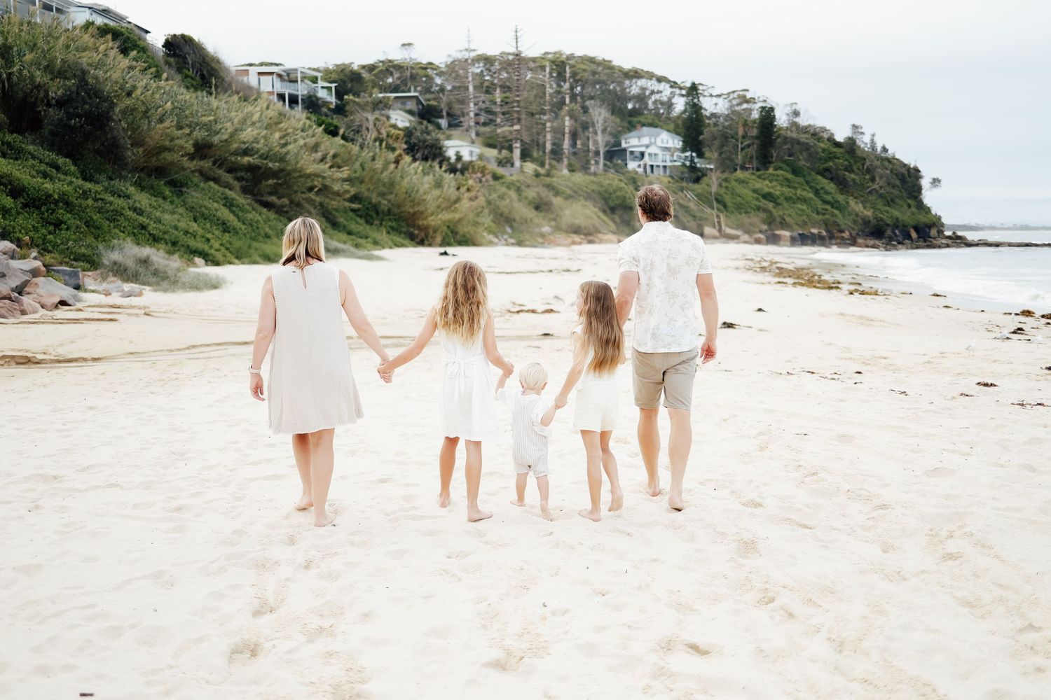 Family of five walking hand-in-hand along a sandy beach toward the ocean.