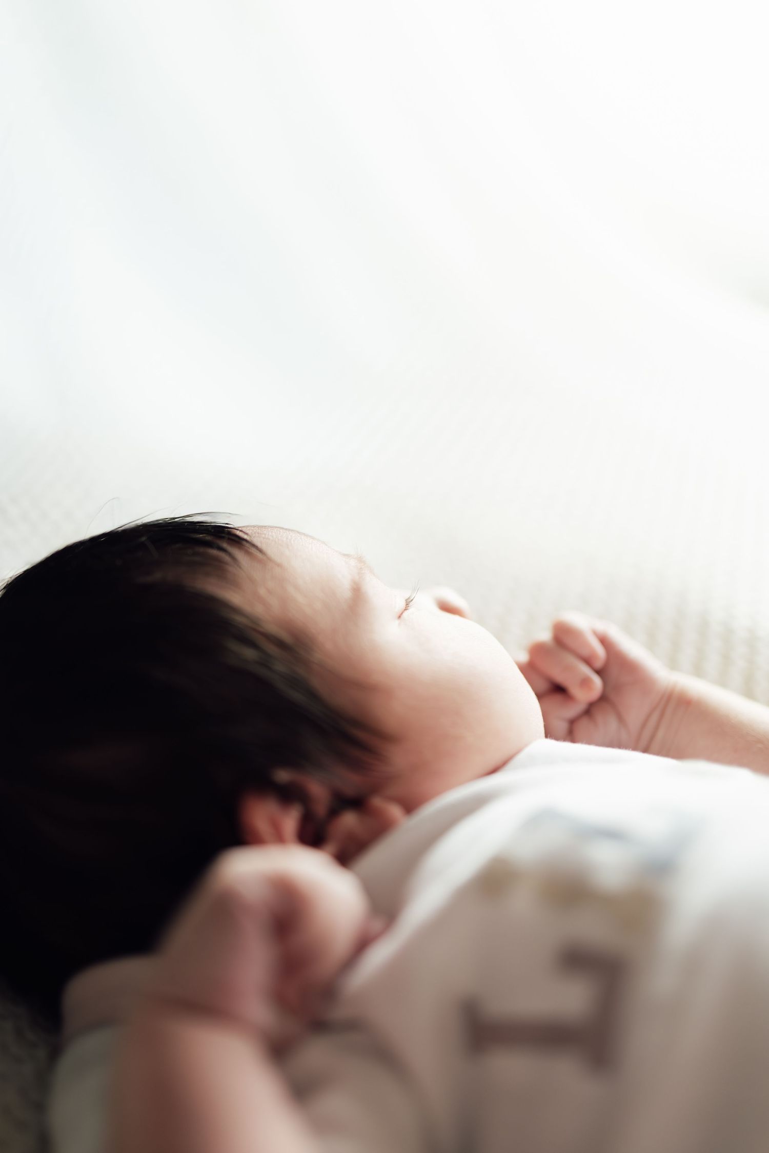A peaceful newborn baby with dark hair sleeping on a white blanket, tiny fists raised beside a serene face.