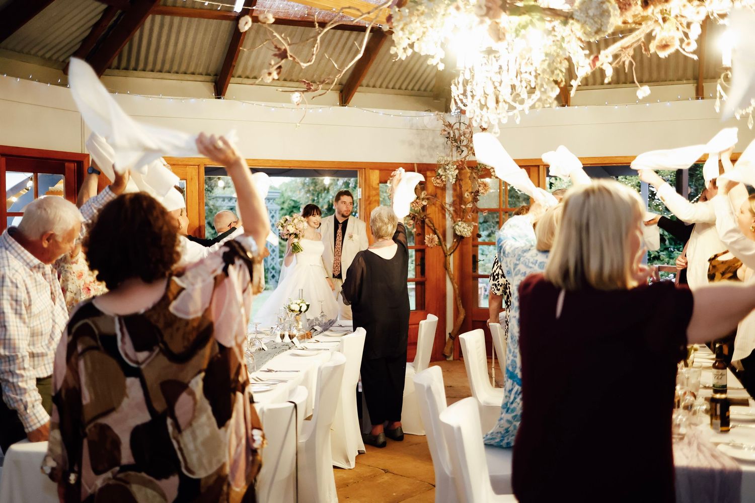 Guests wave white napkins celebrating newlyweds at a romantic rustic barn wedding reception with chandelier lighting.