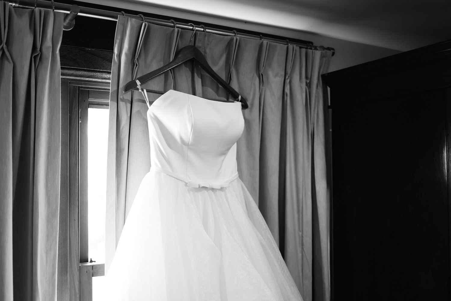 Elegant strapless white wedding ball gown hanging on a hanger against draped curtains in black and white.