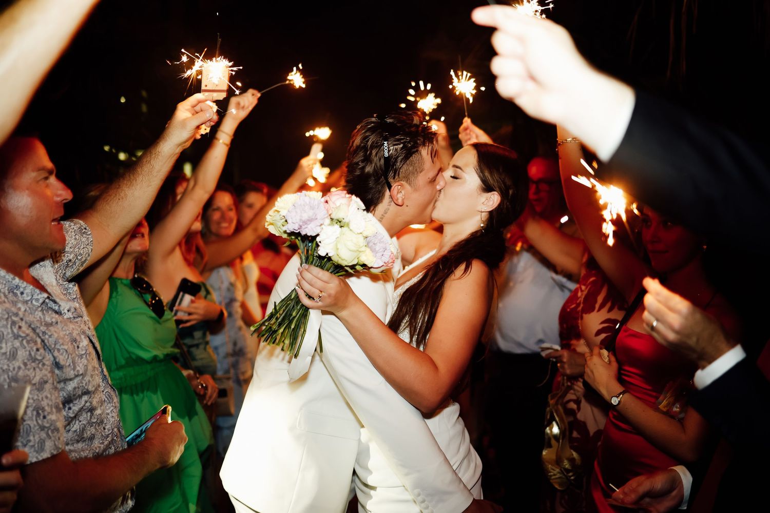 Newlyweds share a romantic kiss surrounded by guests holding sparklers at their evening wedding reception.