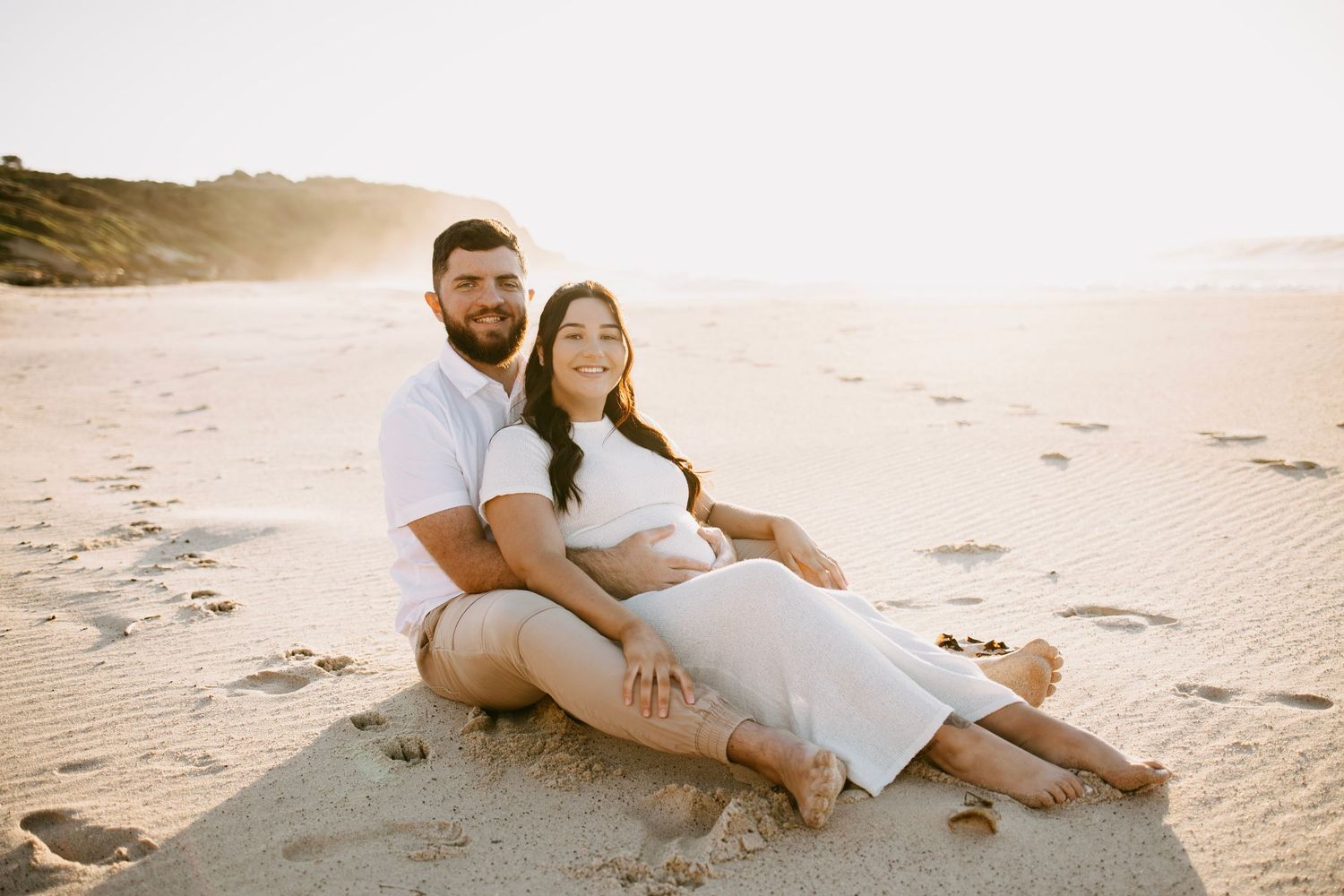Happy couple sitting together on sandy beach at sunset, woman in white dress, man in white shirt.