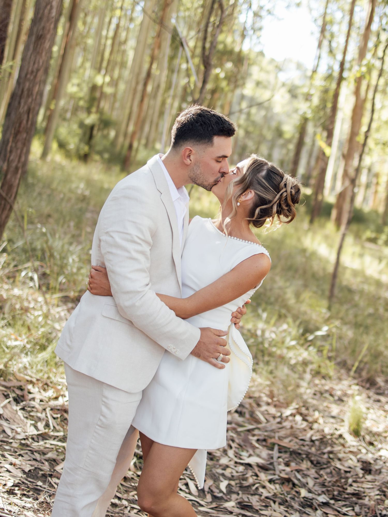 Bride and groom sharing a passionate kiss in sunlit forest wearing elegant white wedding attire.