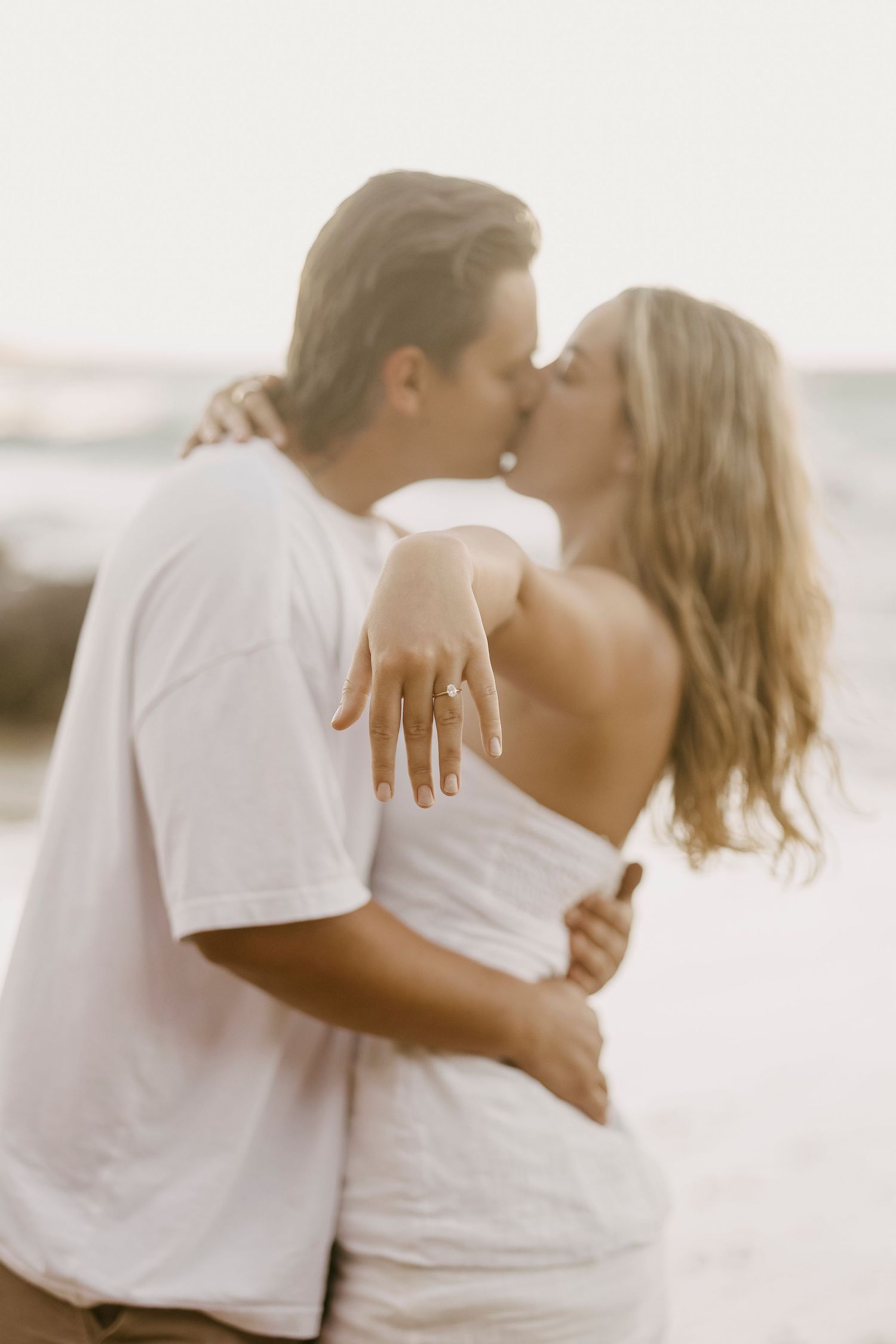 Couple kissing on beach showing off engagement ring at sunset.
