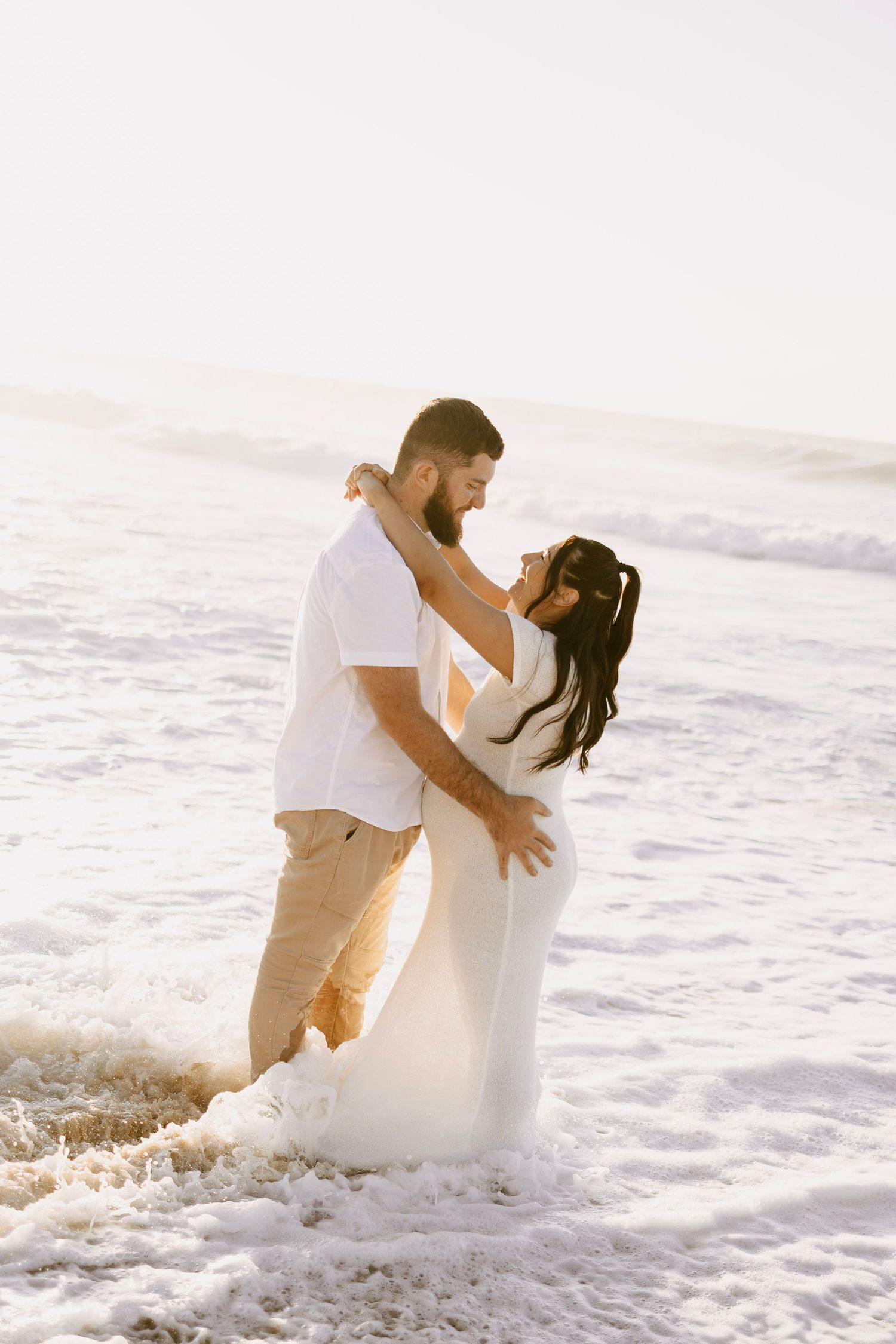 A couple embraces in ocean waves at sunset, the woman wearing a flowing white maternity dress.