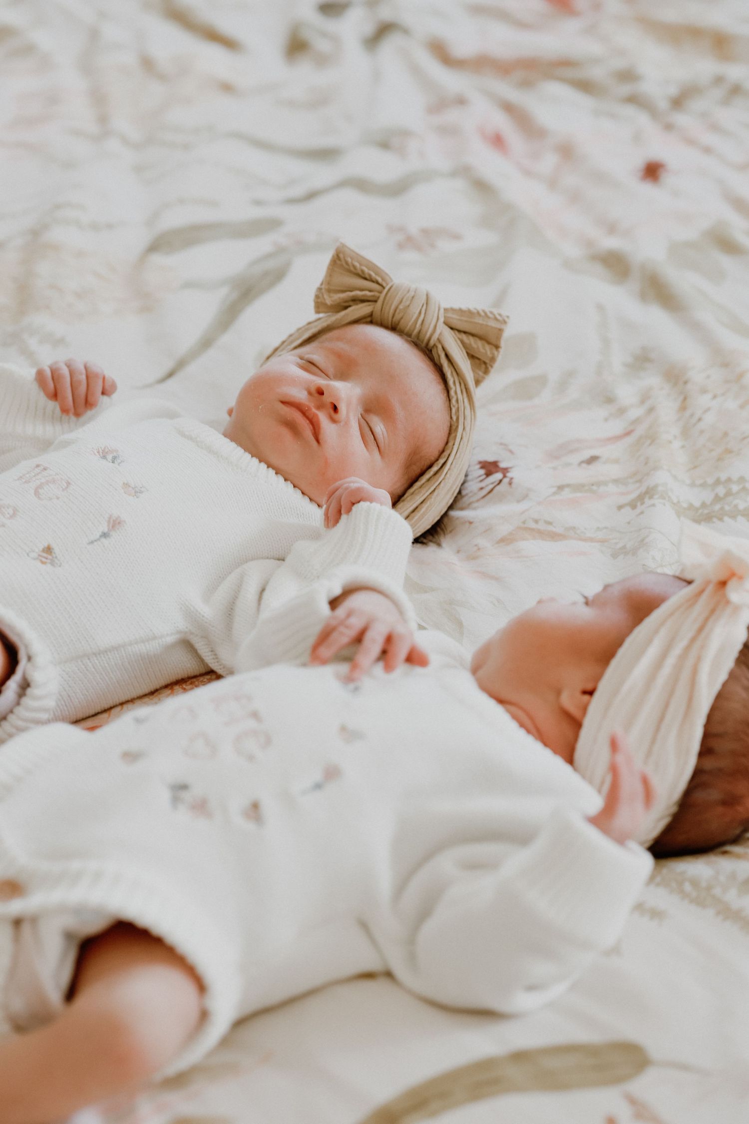 Two sleeping newborn twins in white outfits and bow headbands lying on a soft floral blanket.