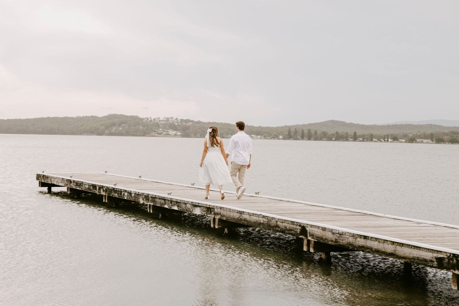 Couple walking hand-in-hand along a wooden pier over calm lake waters on an overcast day.