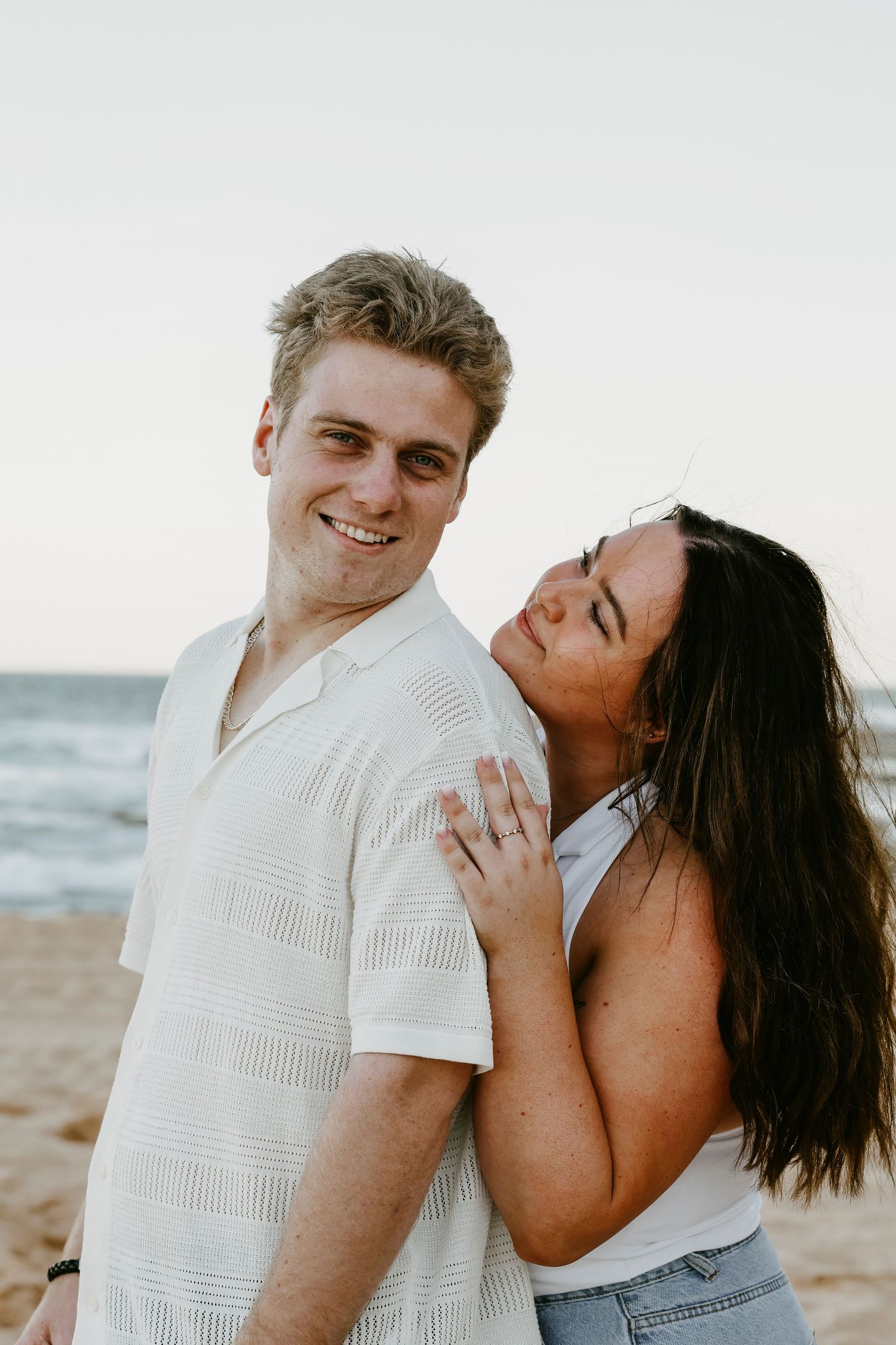 Smiling man and woman posing together on sandy beach, woman leaning against mans shoulder.