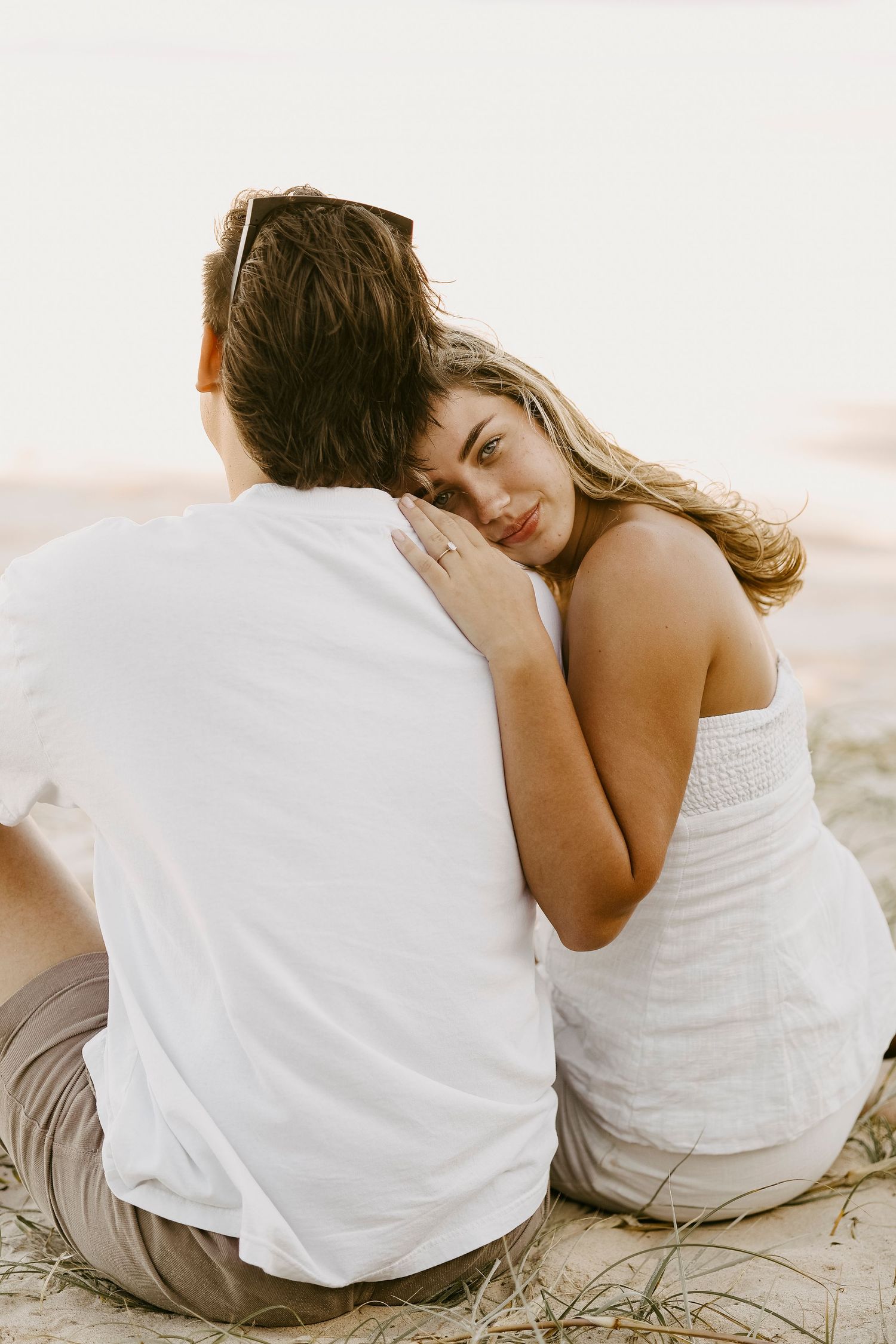 Woman resting her head on her partner's shoulder while sitting together on a sandy beach.