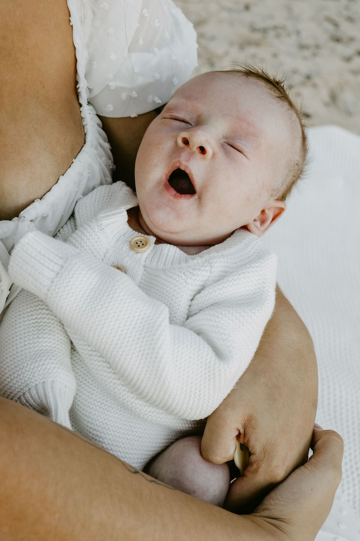 Newborn baby yawning in white knit outfit while cradled in parent's arms during lifestyle photo session.