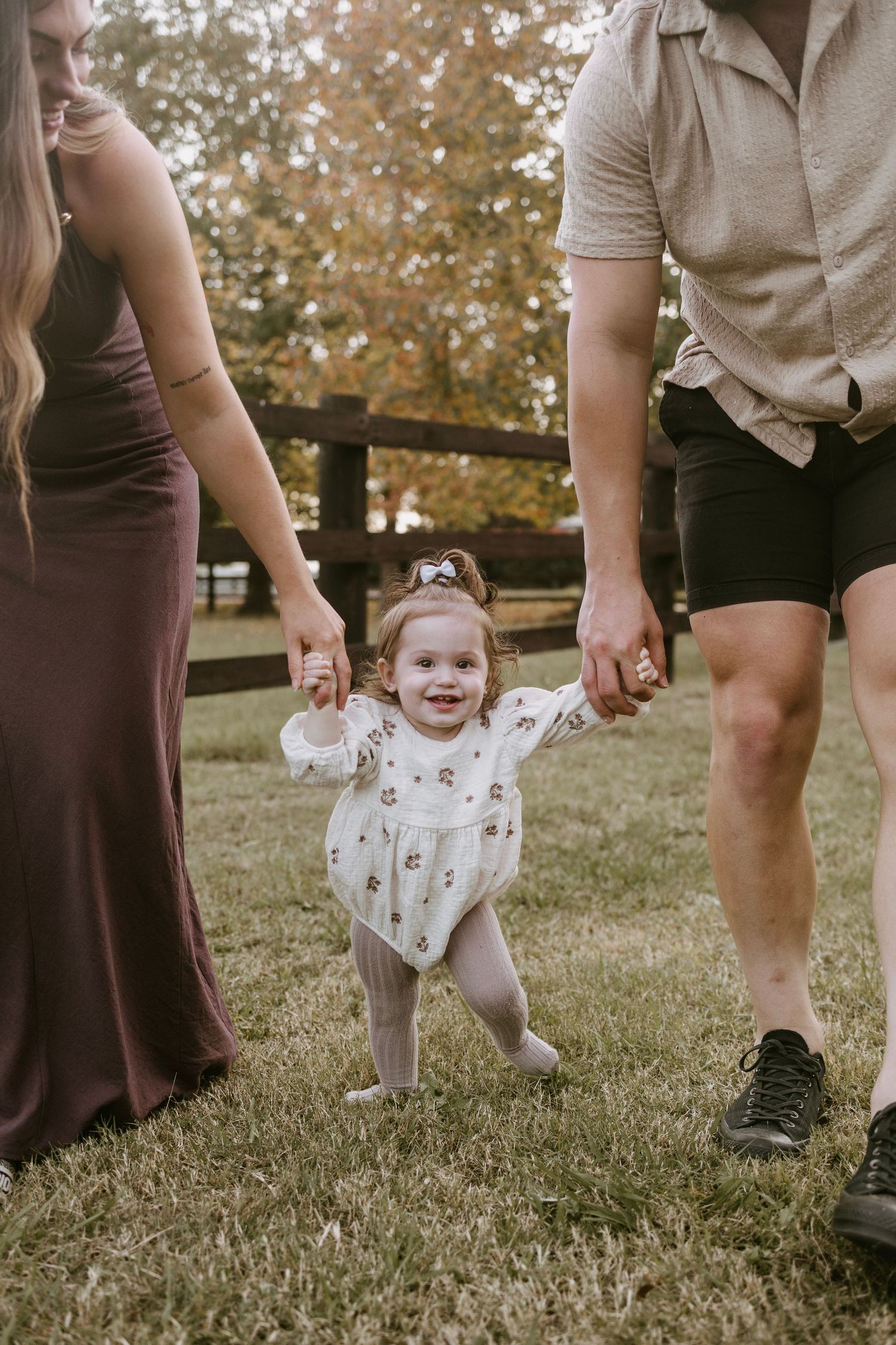 Joyful baby girl taking first steps while parents hold her hands on green grass outdoors.
