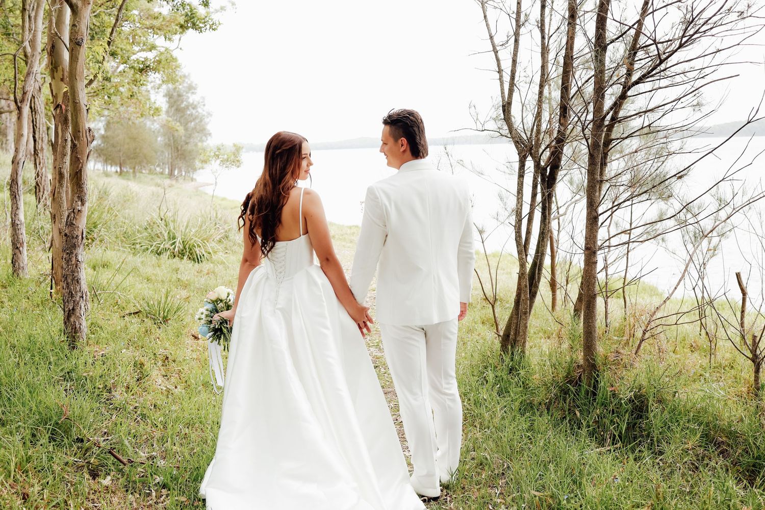 Bride and groom walking hand in hand through a lush green forest path near a lake on their wedding day.