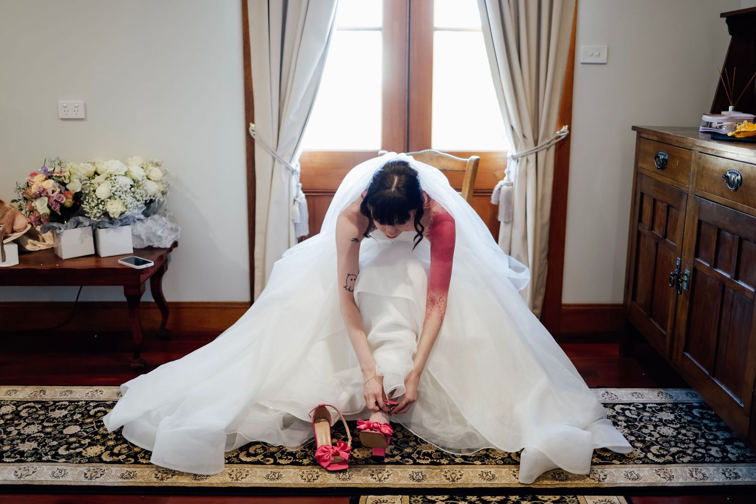 Bride in white wedding gown sitting on floor putting on red shoes near window in elegant room.
