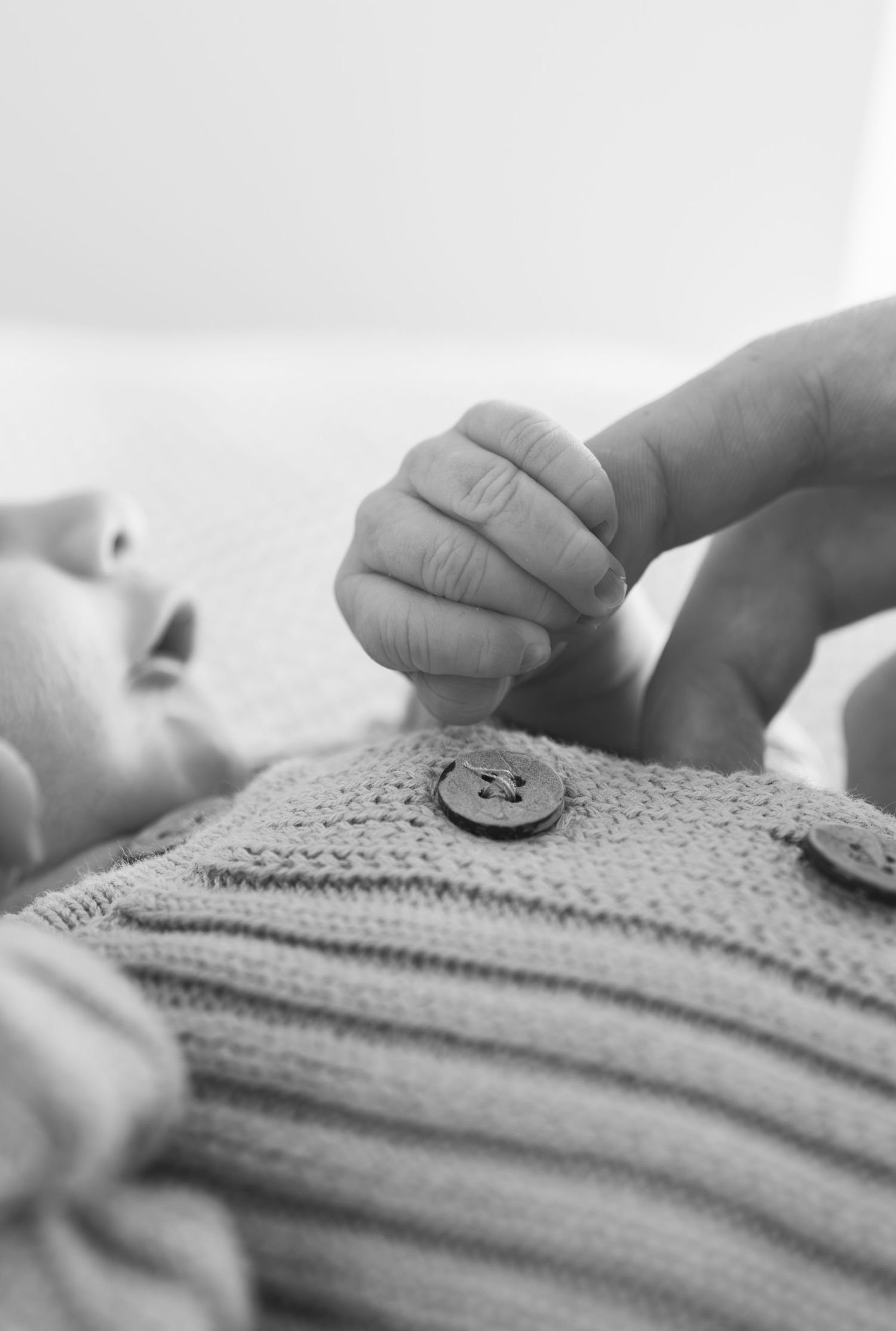 Black and white photo of a newborn baby grasping an adult finger, wearing a knit sweater with buttons.