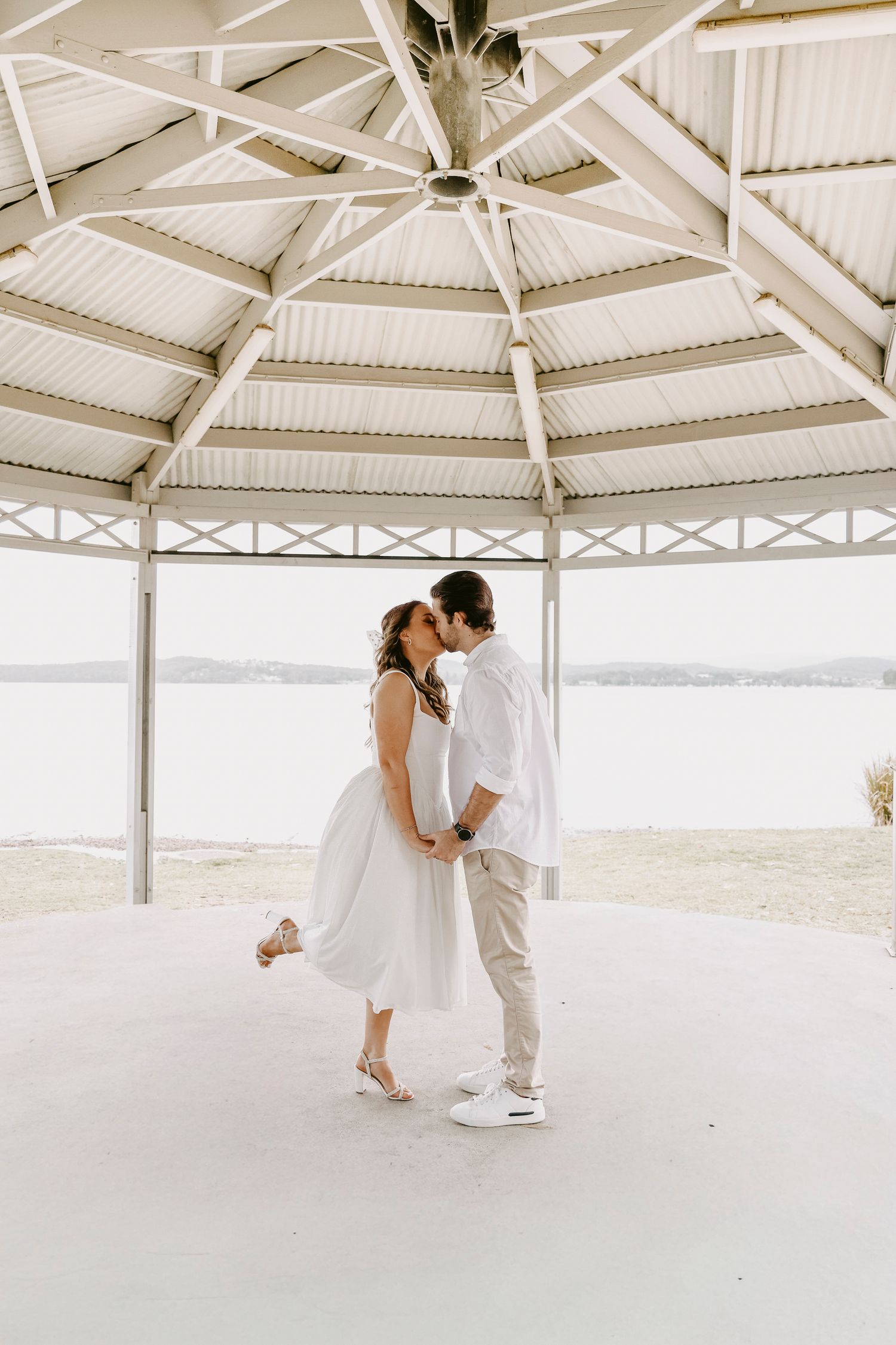 Couple sharing romantic kiss under white pavilion with sandy beach and water in background.