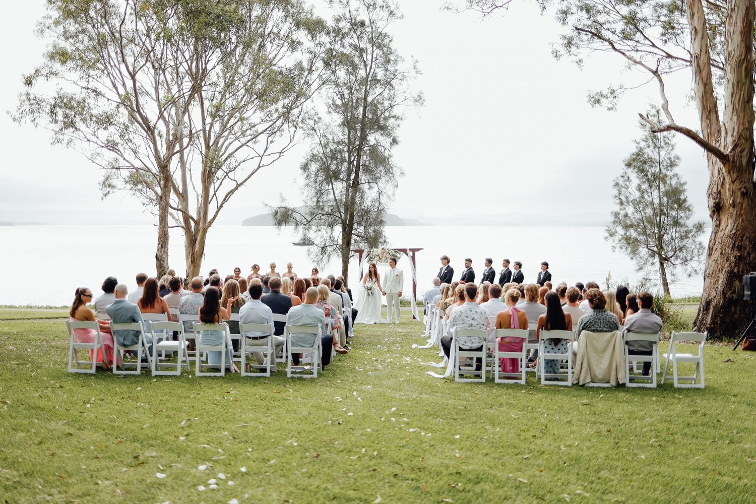 Outdoor wedding ceremony on green lawn by the water with guests seated in white chairs under tall eucalyptus trees.
