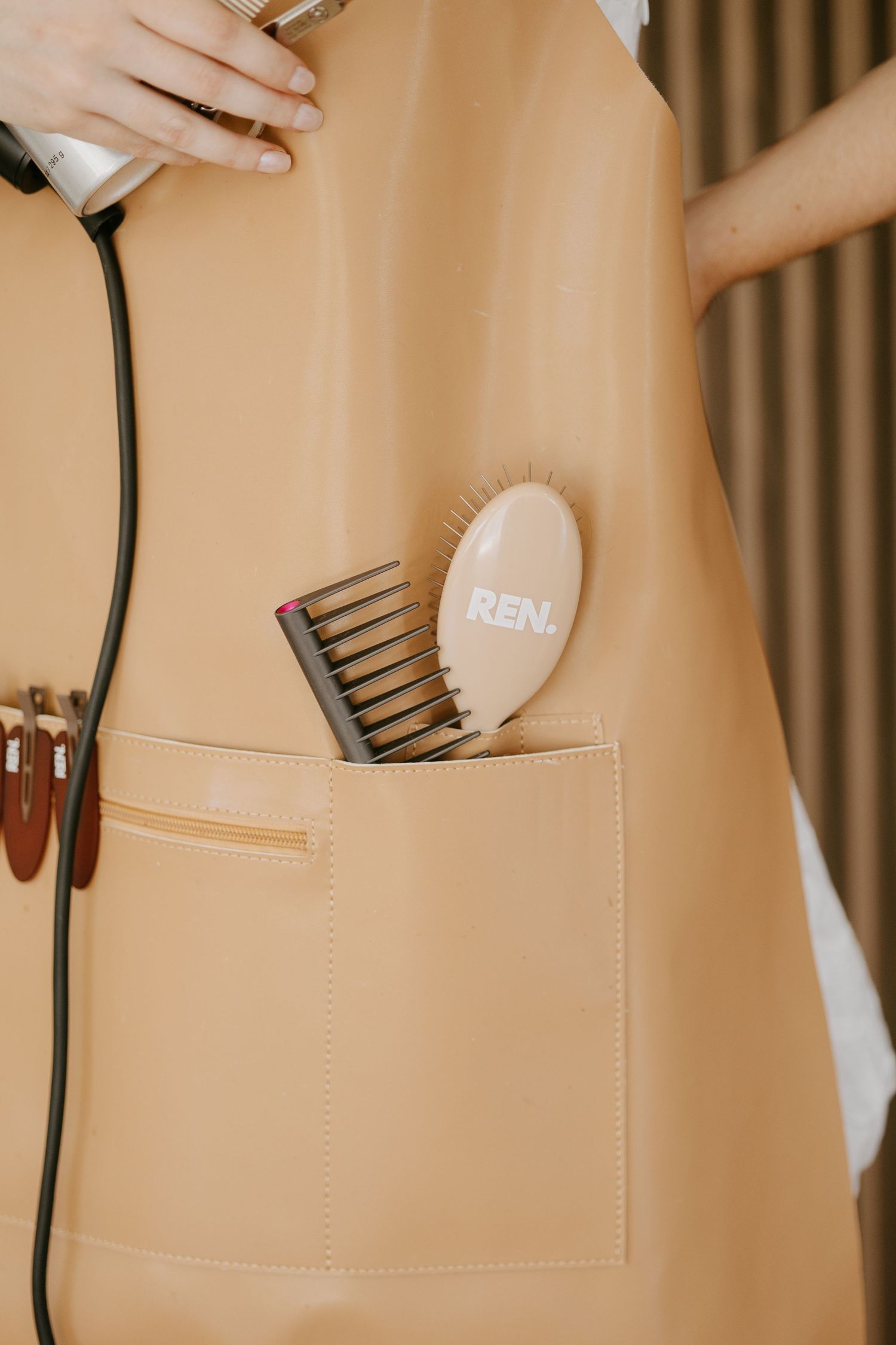 Close-up of a tan salon apron pocket holding a REN hairbrush and comb.