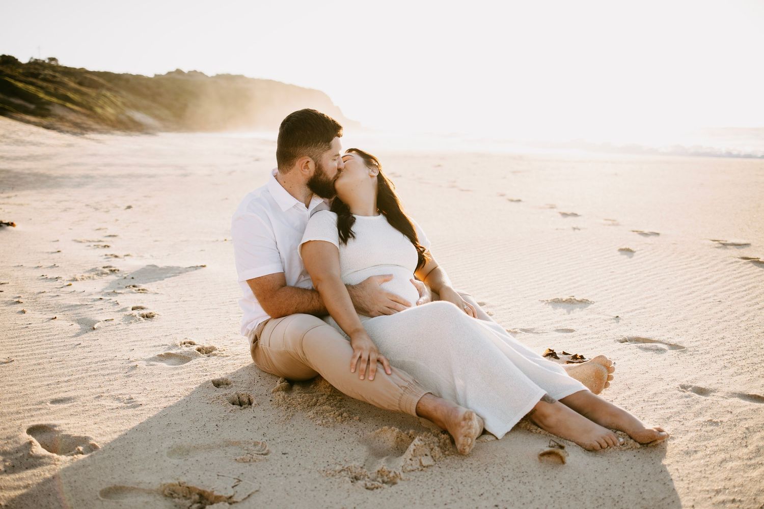 Couple kissing while sitting together on sandy beach at golden sunset hour.