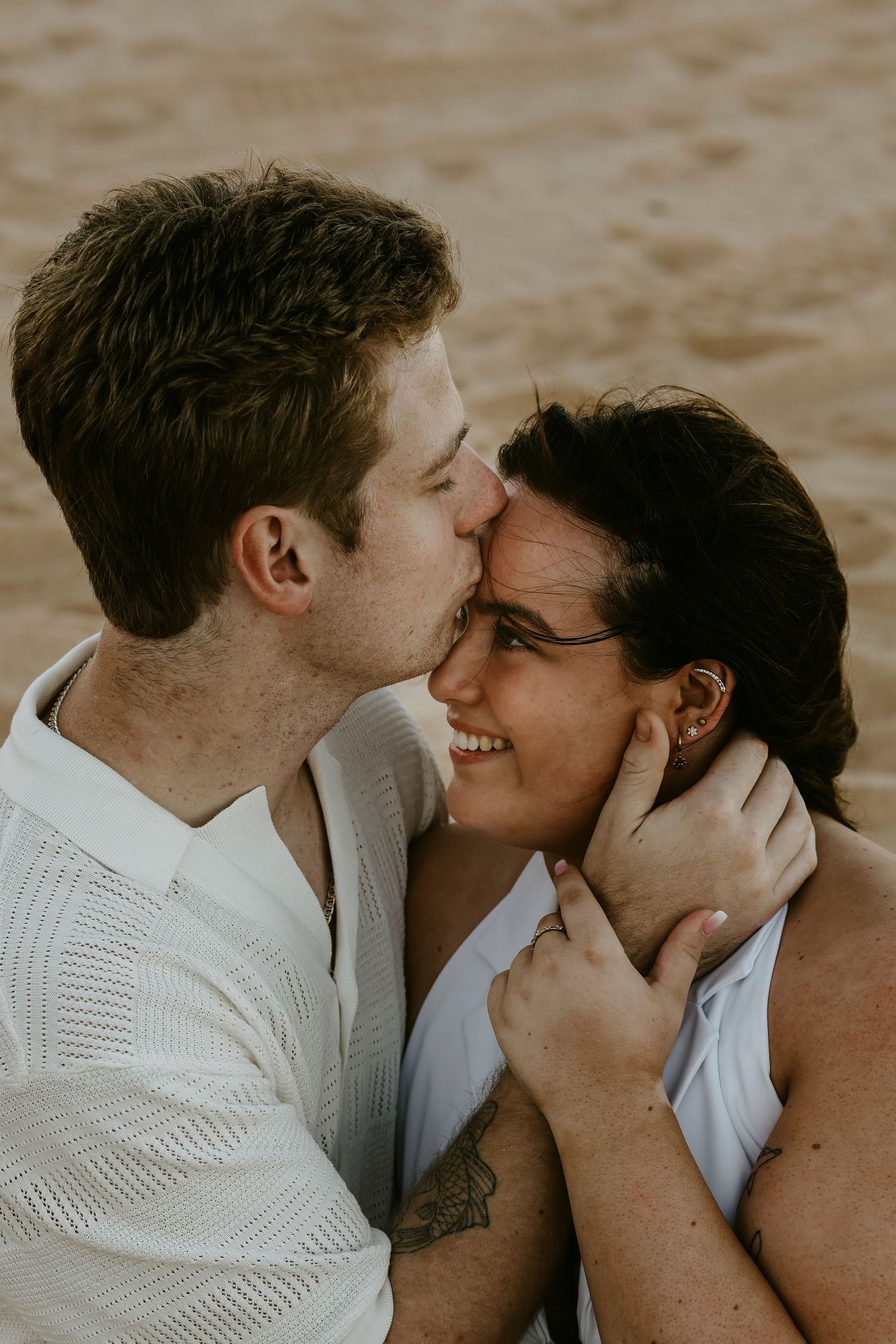 Man gently kissing womans forehead while holding her face on a sandy beach.