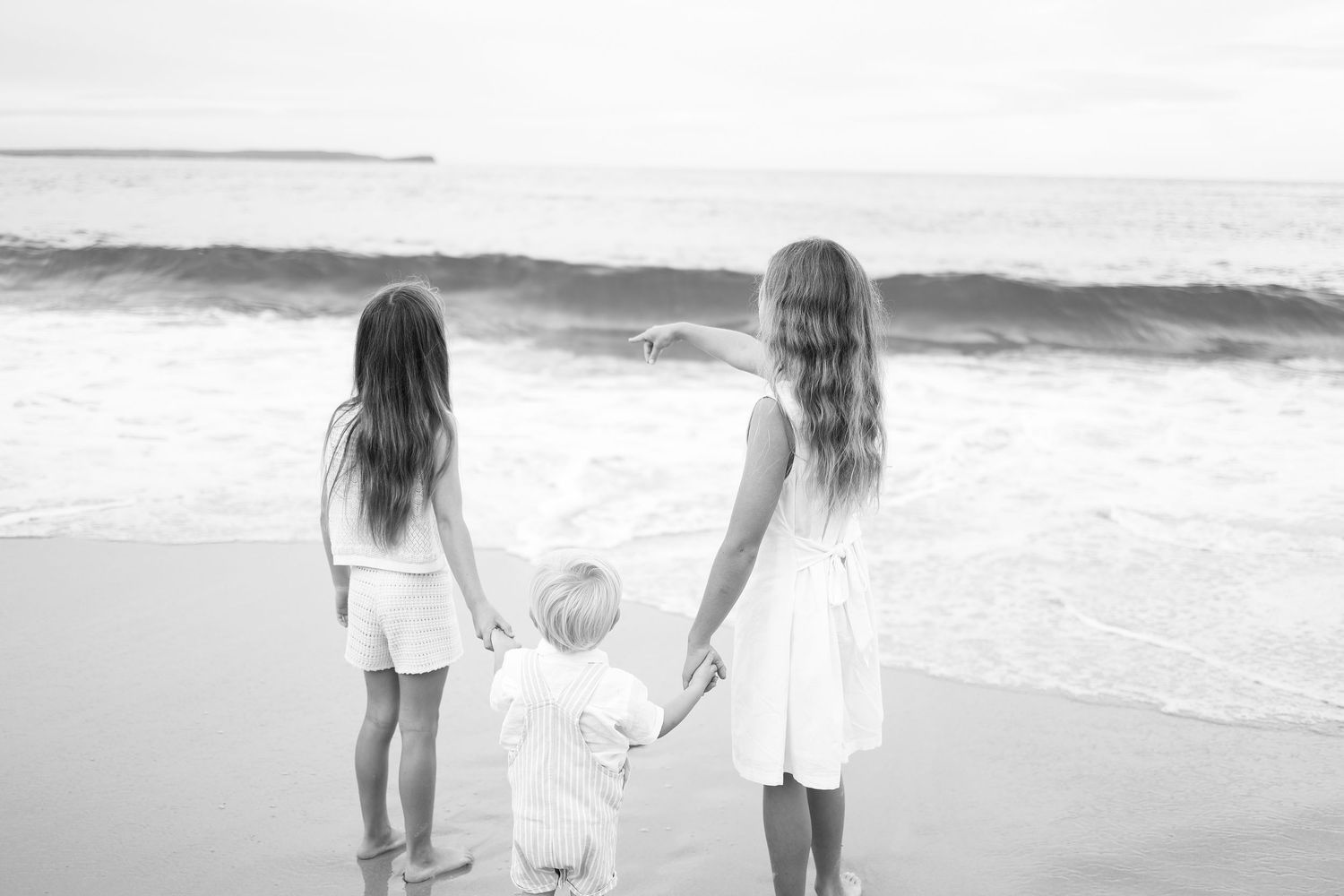 Black and white image of two girls and a toddler holding hands facing ocean waves.