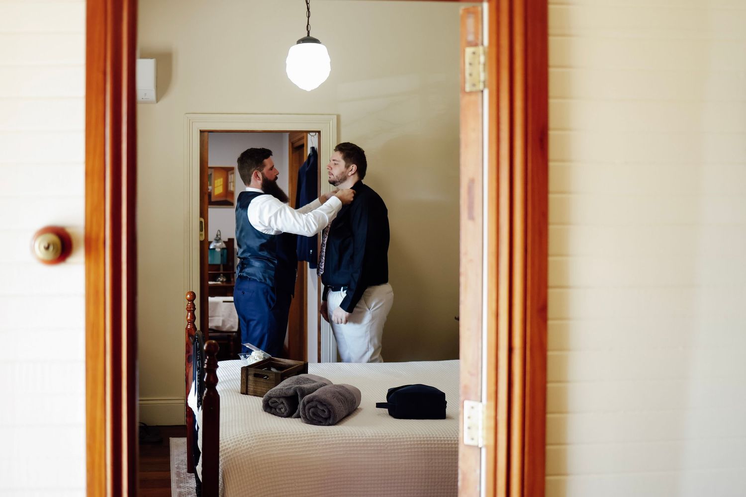 Two men helping each other get dressed in a warmly lit bedroom before a wedding ceremony.