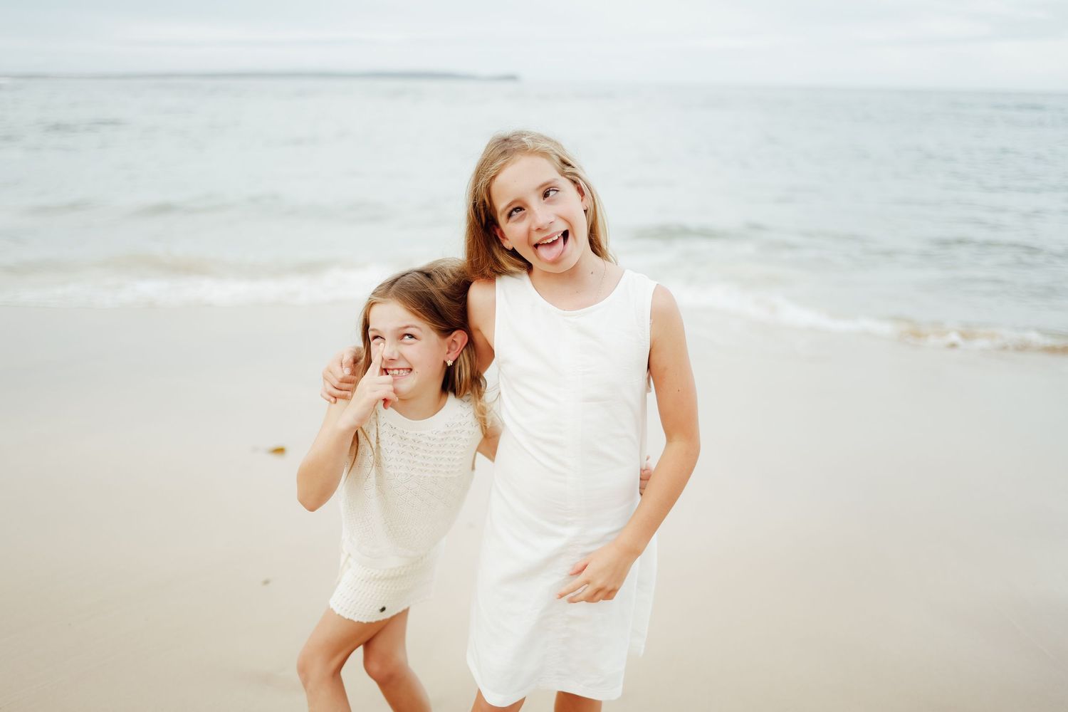 Two happy sisters in white dresses laughing and hugging on ocean beach shoreline.