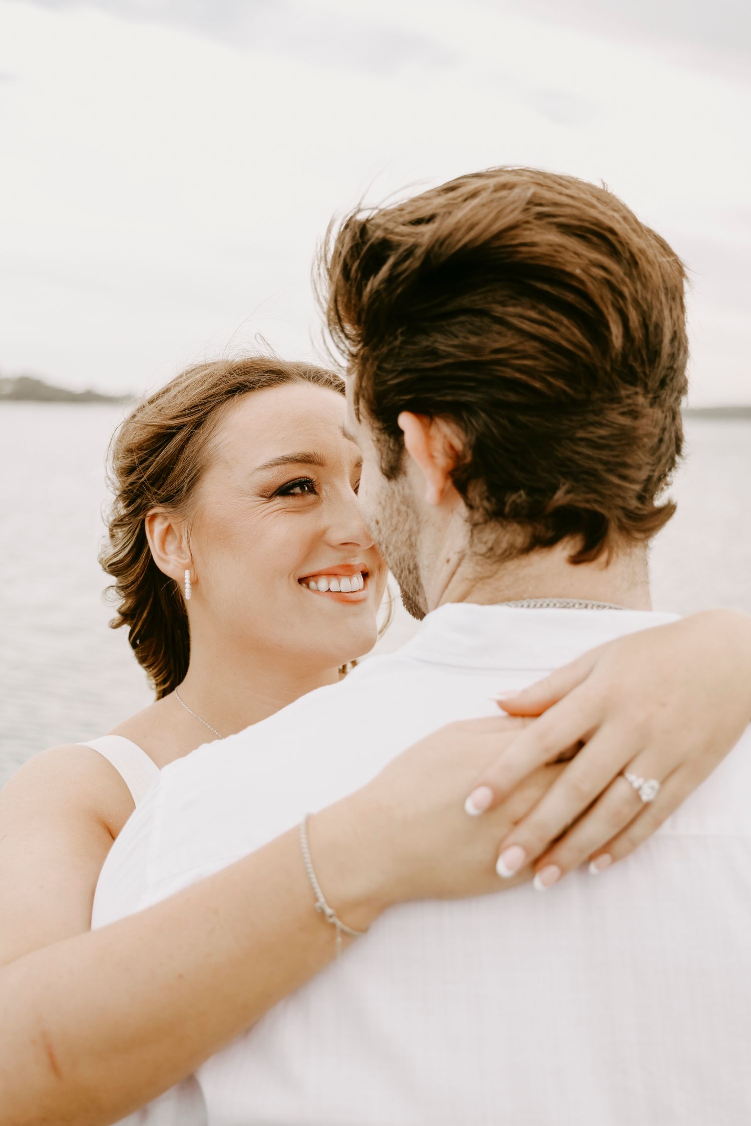 Smiling bride-to-be embracing her partner near water, showcasing her engagement ring.